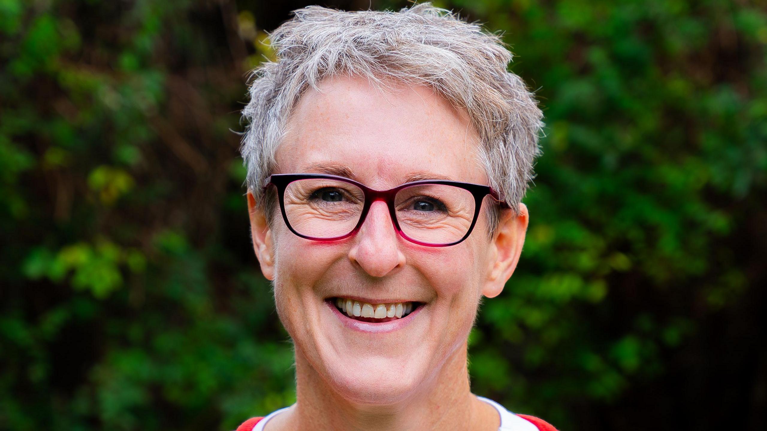 Sarah Haythornwaite - a woman with grey hair wearing purple glasses, smiling for the camera, with trees in the background.