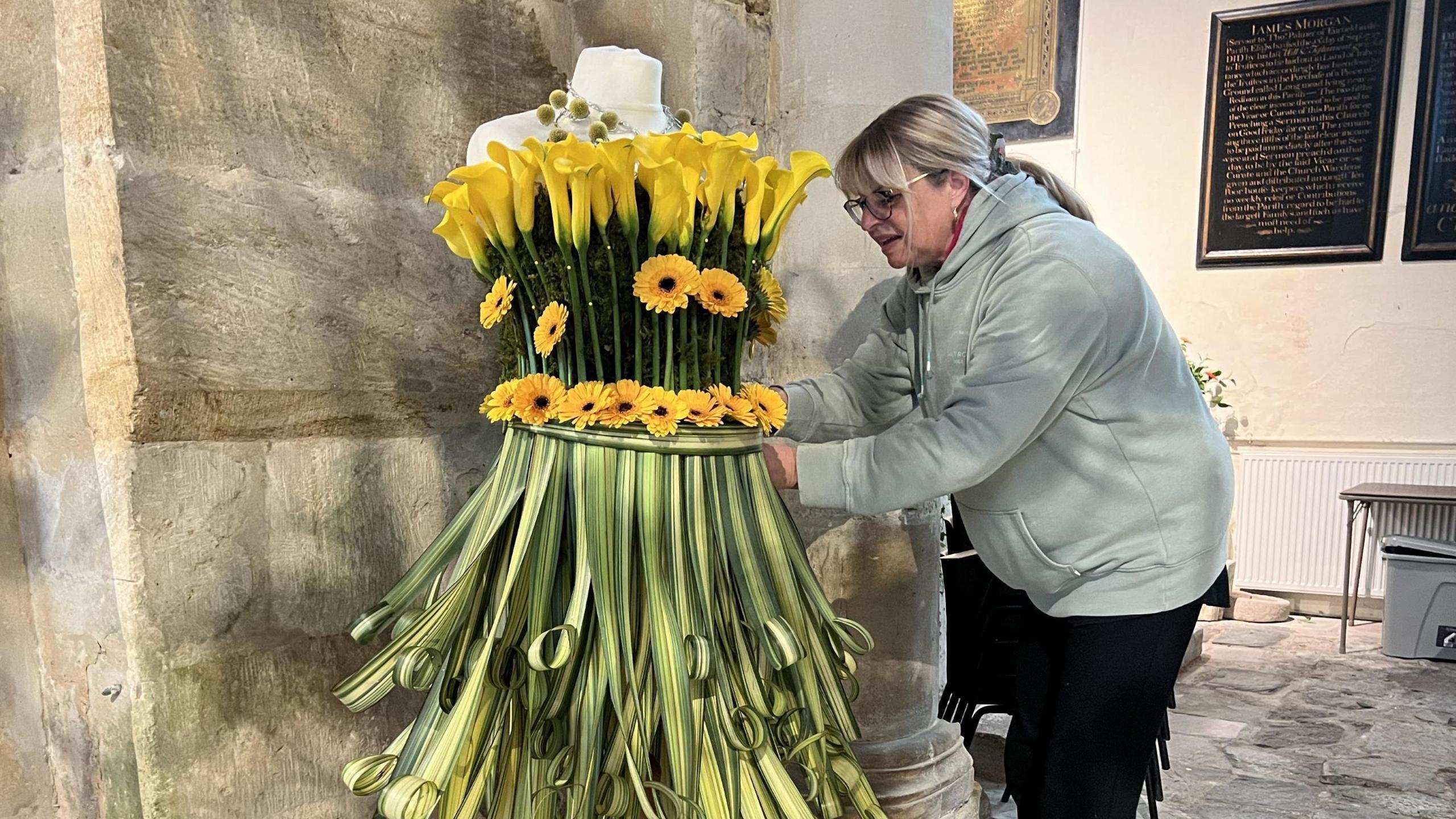 A woman with blonde hair wearing a grey top and black trousers tends to an elaborate flower display inside St Andrew's in Stogursey in Somerset. The display is of largely green and yellow blooms