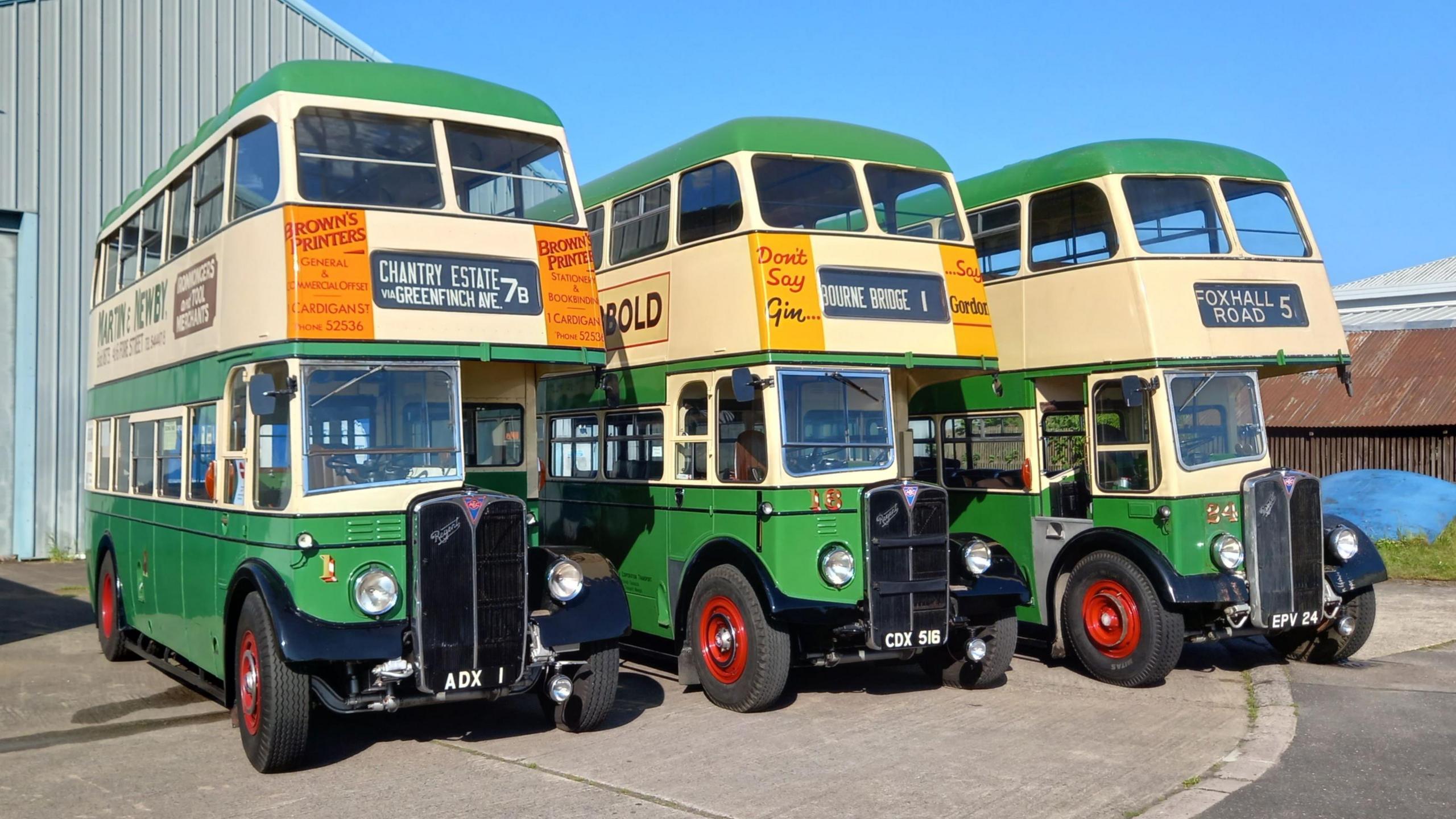 The three classic double decker buses are lined up outside a barn. The sky is blue and the buses are painted green and white, while the wheels are bright red.