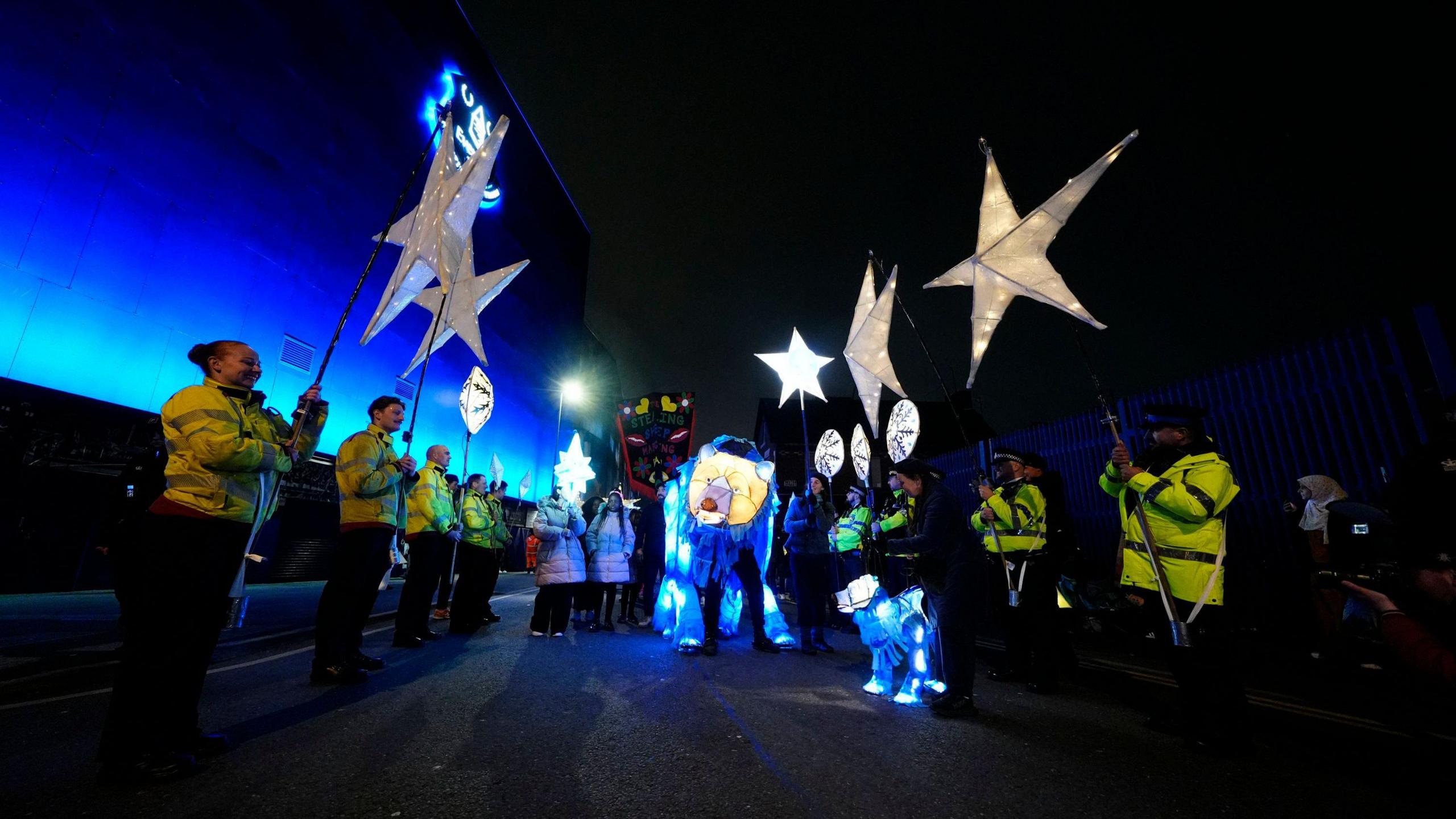 Staff wear hi-vis jackets holding lit up star lanterns, with the bear lantern in the middle of the crowd 