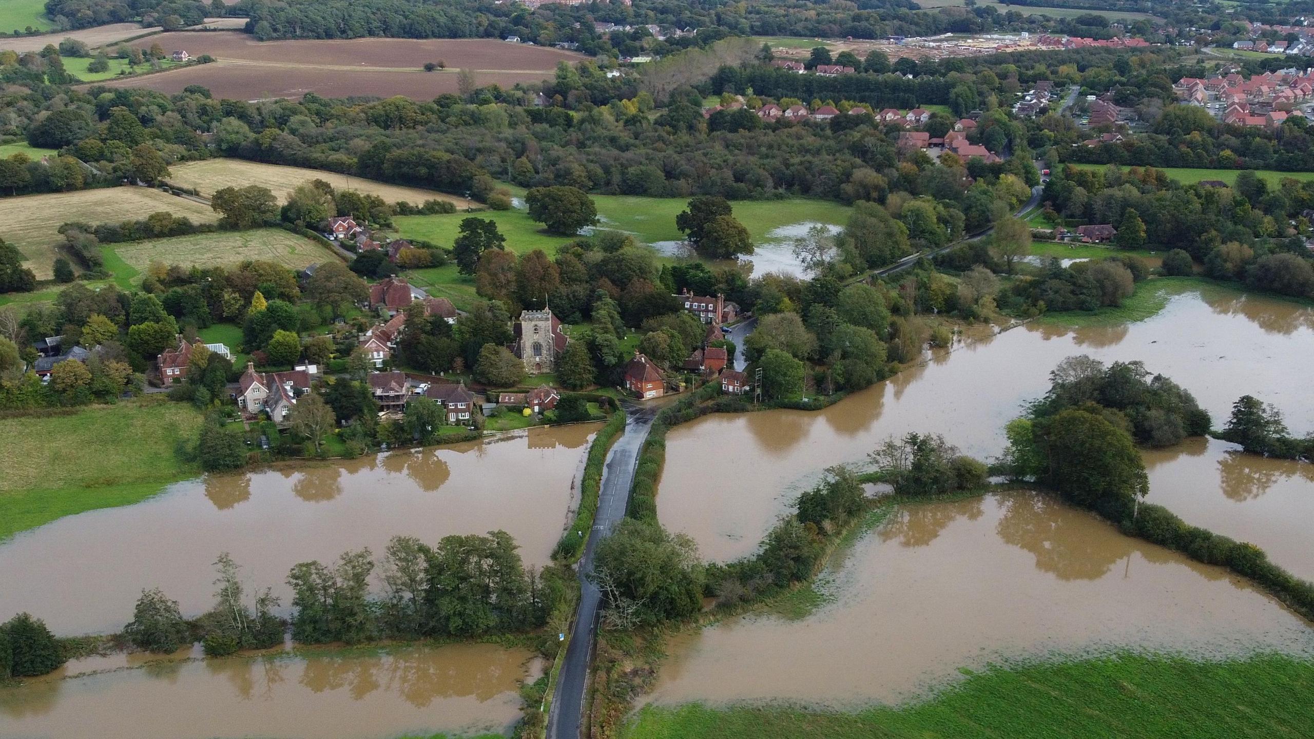 An aerial view showing a flooded Sussex village with fields in the foreground that are completely underwater, a church and a few houses around it are also seen while in the background there are more homes, tree and other fields, some with flooding.