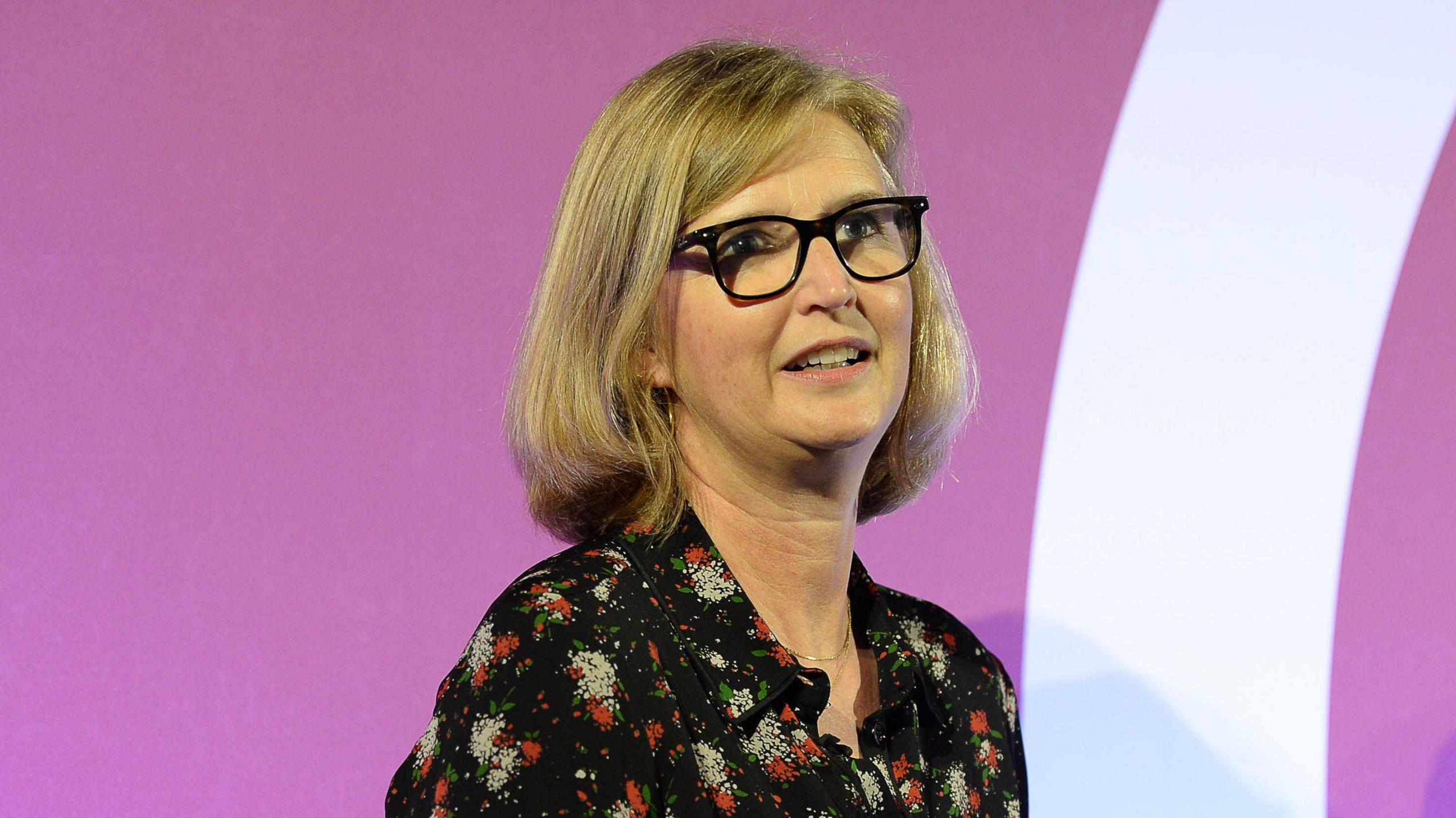 Side profile of Joanna Prior seated with glasses, blonde bob, wearing dark patterned dress. Against purple backdrop