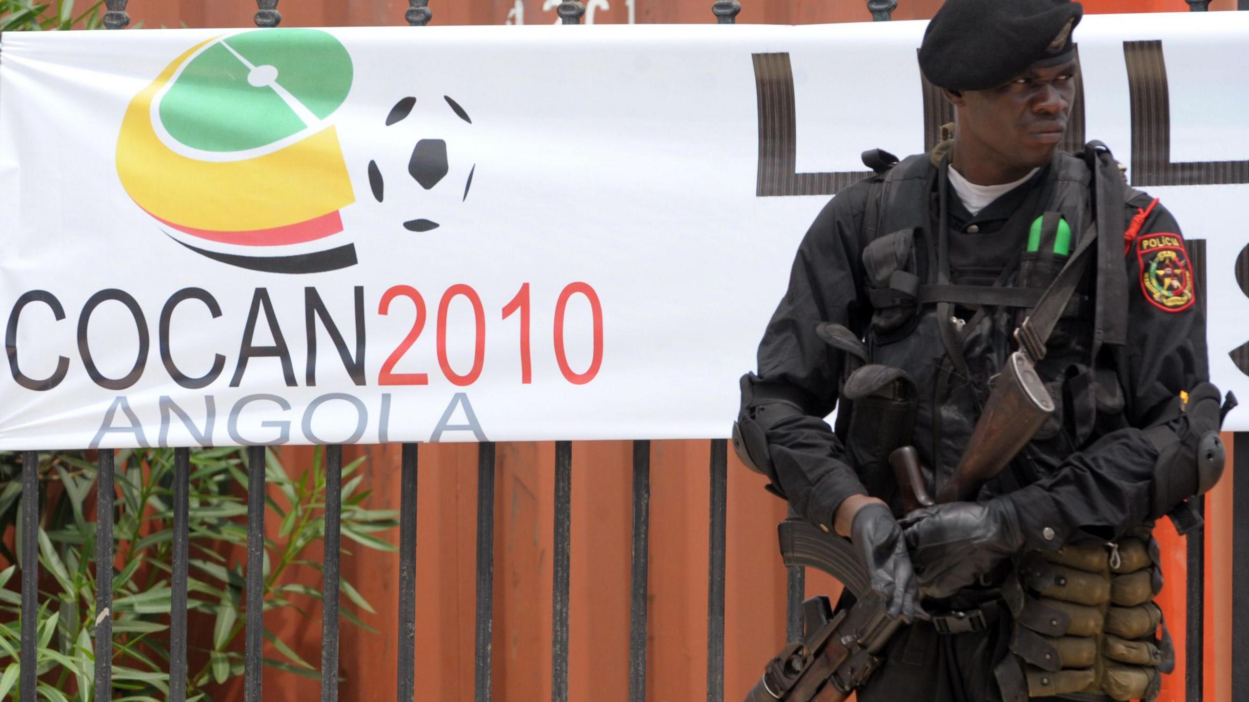 An Angolan policeman, dressed in all black with a beret and automatic rifle, stands guard in front of iron railings which bear a banner with the 2010 Africa Cup of Nations logo on it