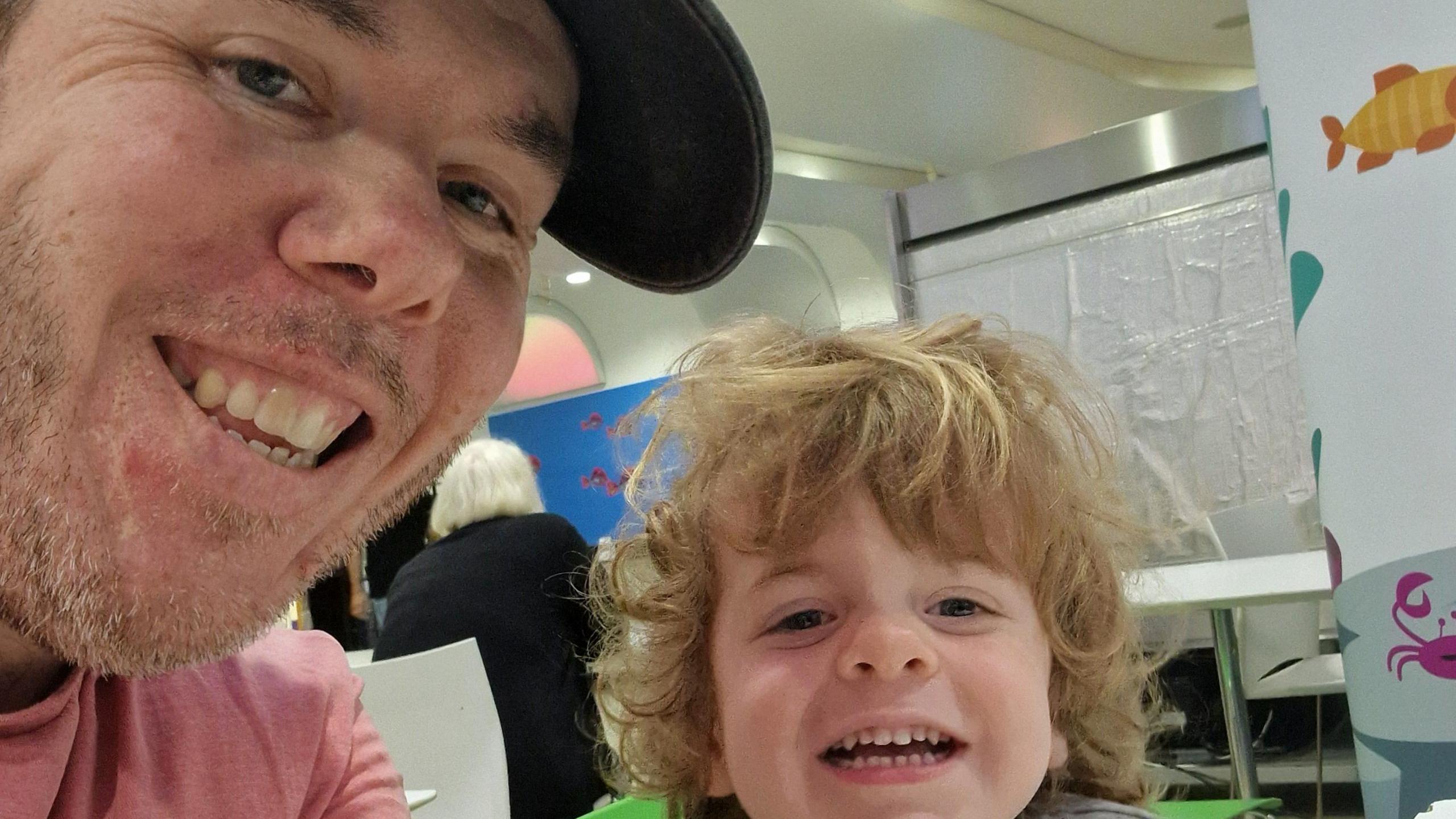 A close up photo of David Bedwell with his son Wilfred. They are both smiling to the camera. David is wearing a cap and a pink top. He has a stubble beard. Wilfred's head can be seen next to him. A hospital setting can be seen behind them.