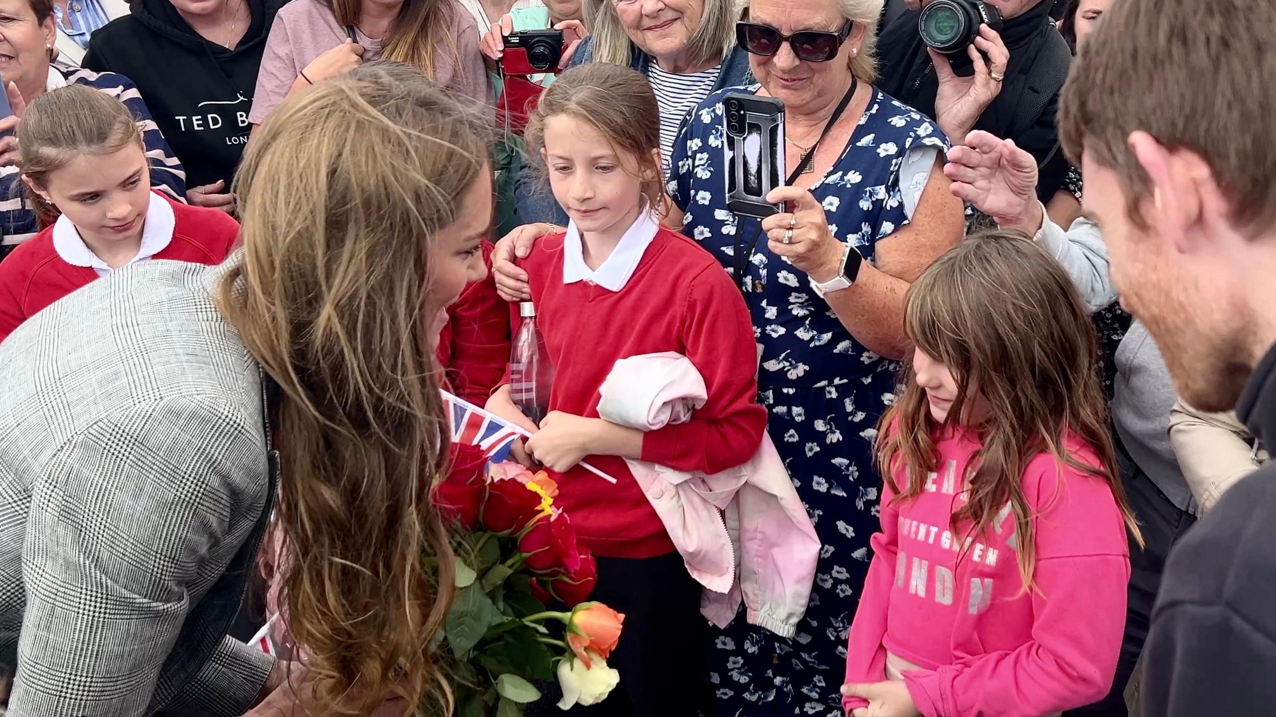 The Princess of Wales kneels down slightly as she speaks to a young girl standing at the front of a crowd. The girl has long brown hair and is wearing a pink hoodie.
