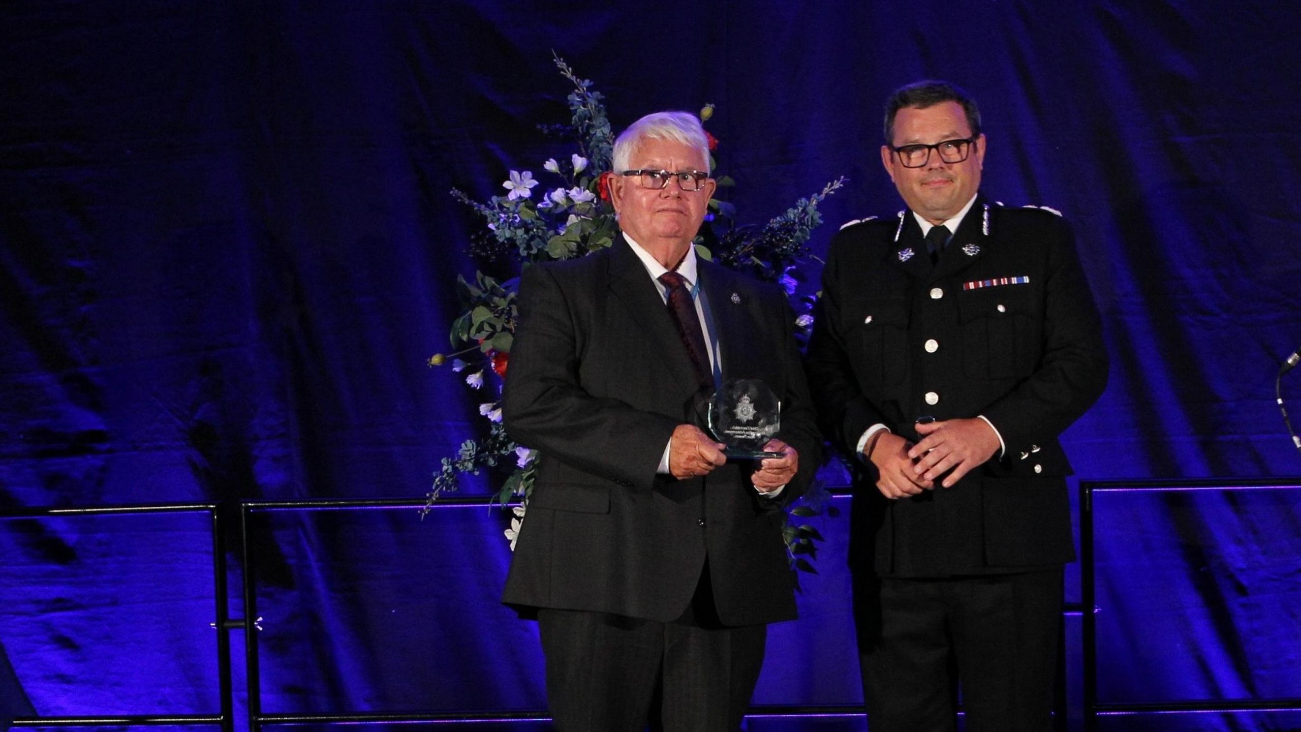 Glan Thomas holding his trophy on the stage alongside a sergeant on stage. Both are looking at the camera. Behind them is a large flower arrangement. Glan, on the left, is wearing a black and white suit with a maroon tie. On the right, the sergeant is wearing a black officer's suit with a white shirt and black tie.