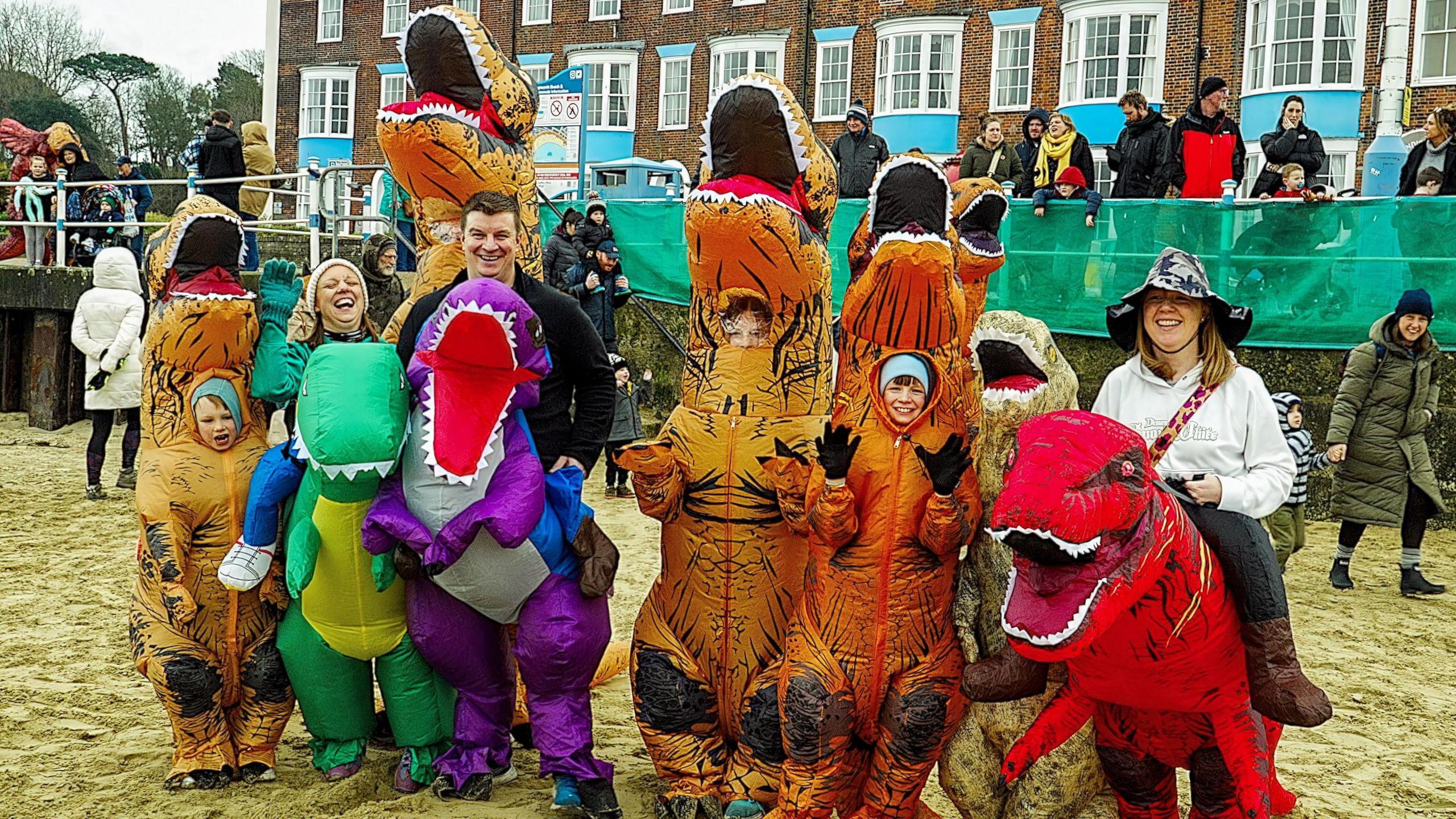 Three adults and three children, all dressed in dinosaur costumes, standing in a row on a beach and smiling at the camera.