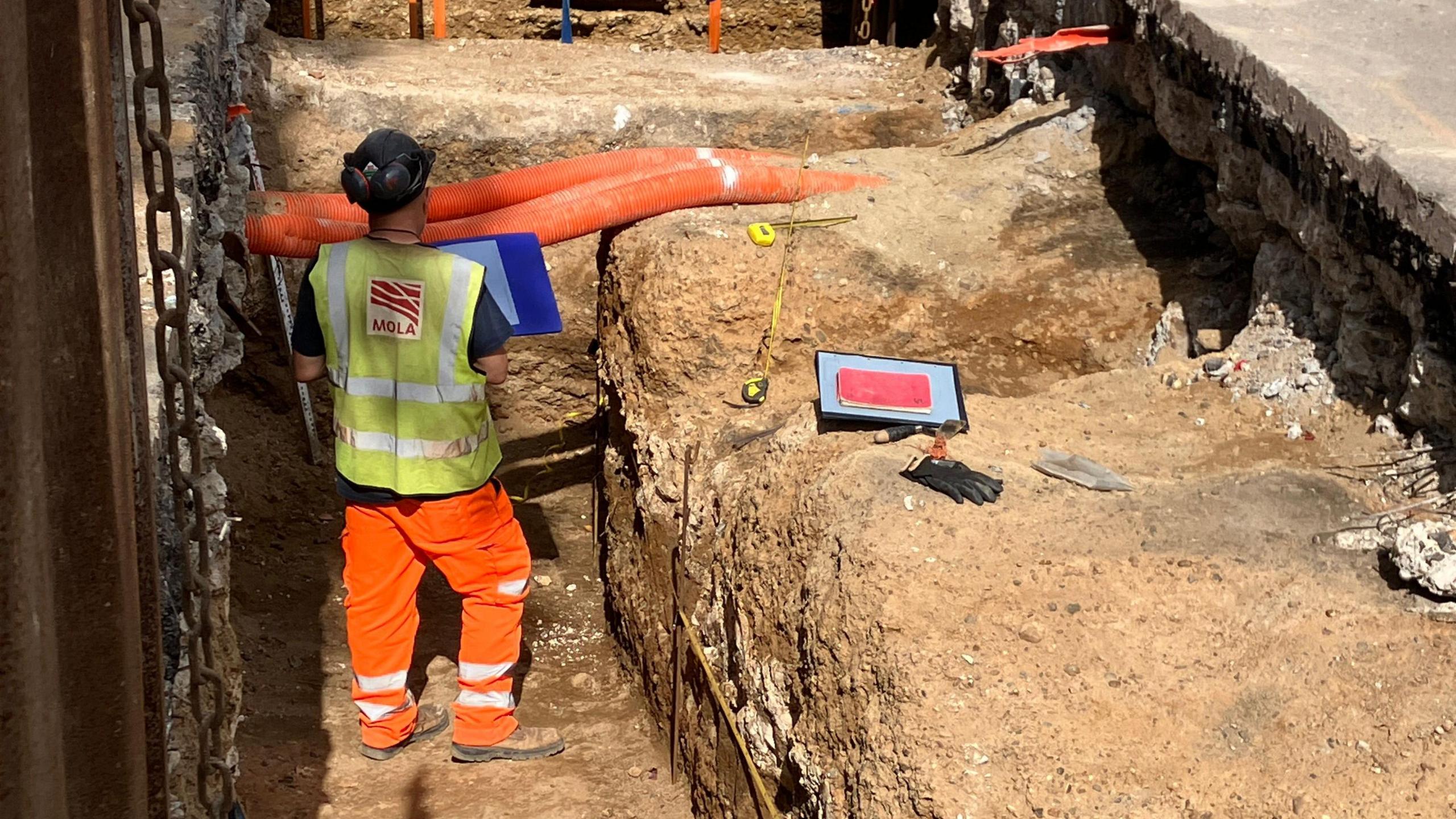 A man wearing a black hard hat and yellow and orange hi vis clothing stands in an excavated pit next to the section of Roman road. Yellow tape measures, a red notebook, blue folder and black gloves are visible on the ground next to him, with orange tubes in the background