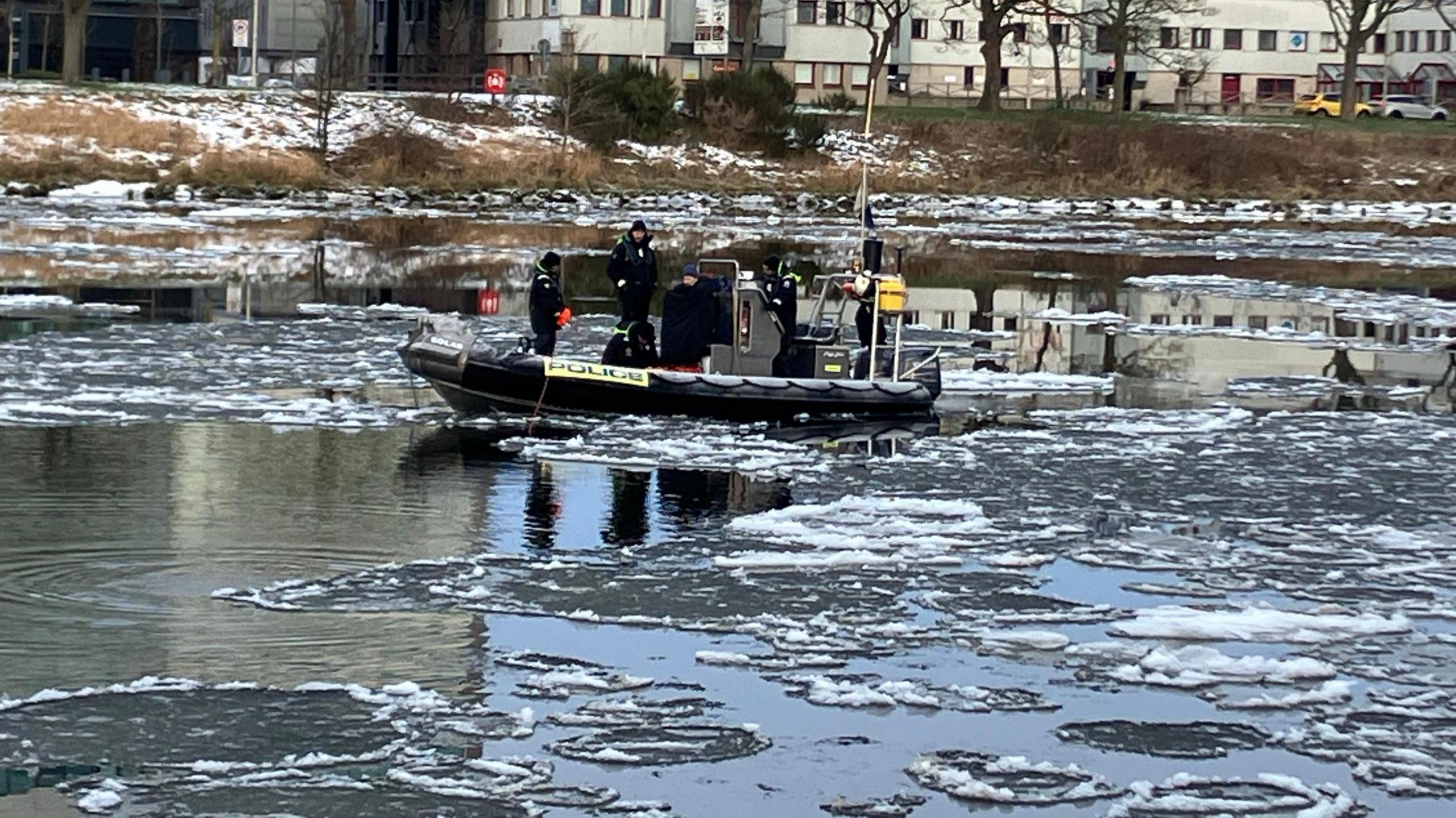 A black Police boat surrounded by patches of ice on the River Dee. Officers dressed in black clothing can be seen on board. The river bank is partially covered by snow.