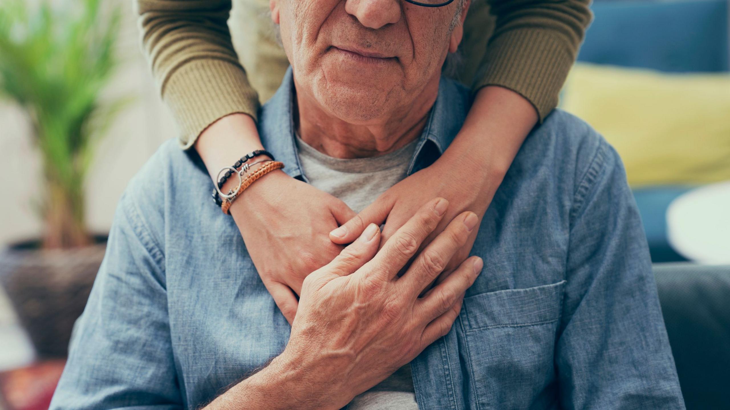 A man wearing a blue shirt with a woman, wearing an olive green top, with her arms draped over his shoulders. The two people are holding hands.