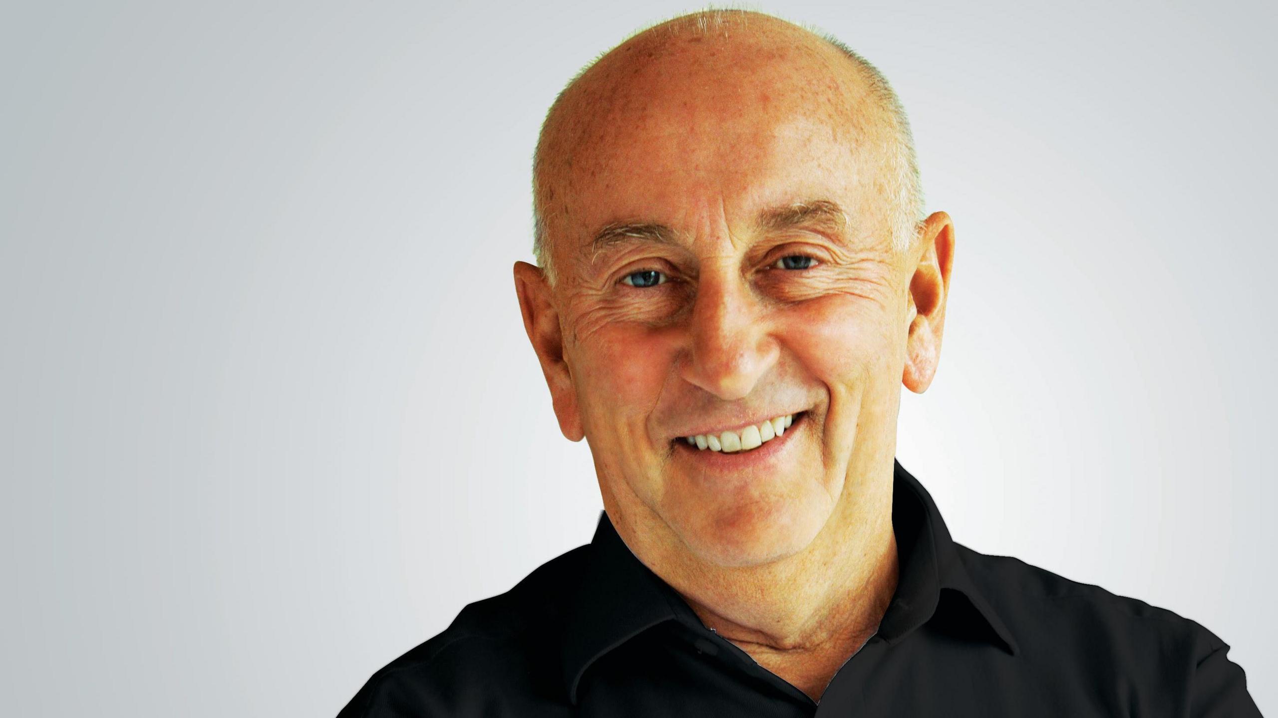 A smiling Fred Done, with a bald head and wearing a black collared shirt, stands in front of a light grey background