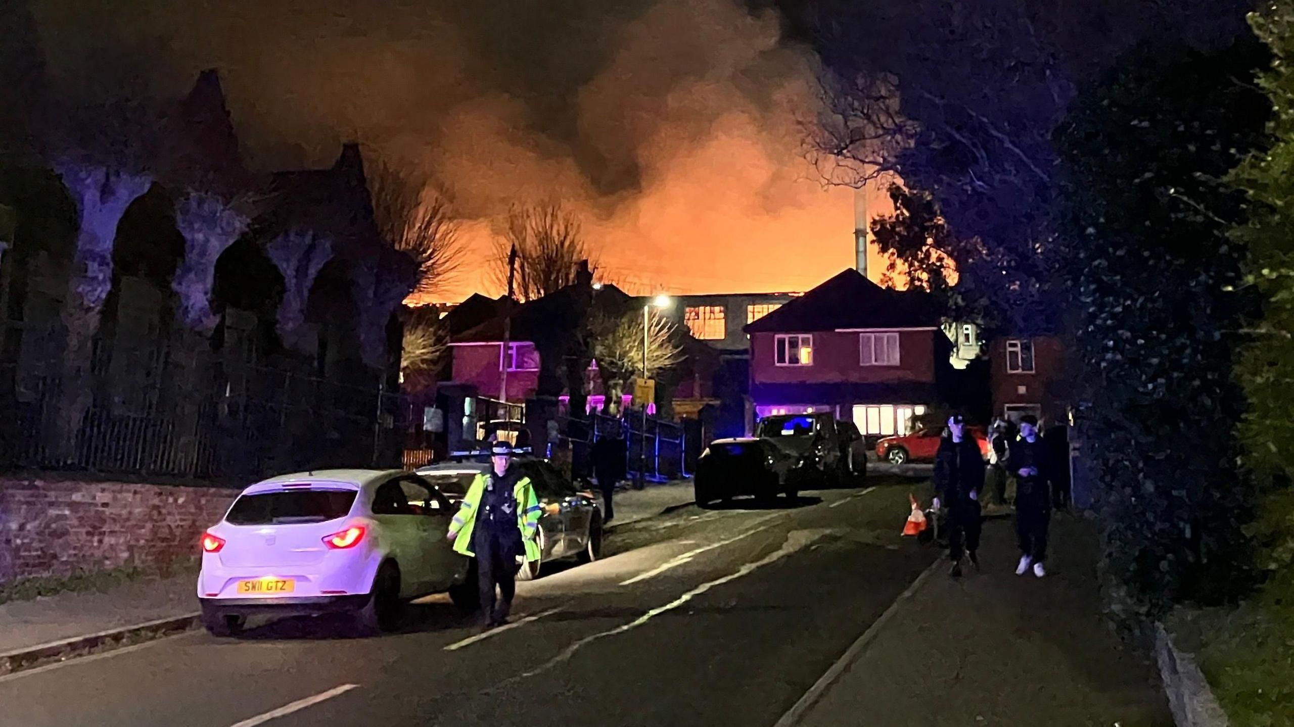 Smoke and an orange glow can be seen coming from behind residential home. There is a police officer and a car in the picture