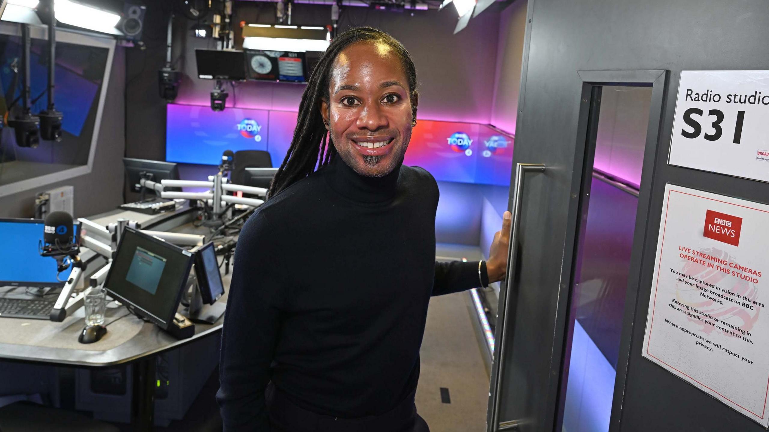 Headshot showing Professor Jason Arday looking into the camera smiling,  standing inside the BBC's Today programme studio. He is a black man with long black locs, ear piercings, wearing a black polo neck.