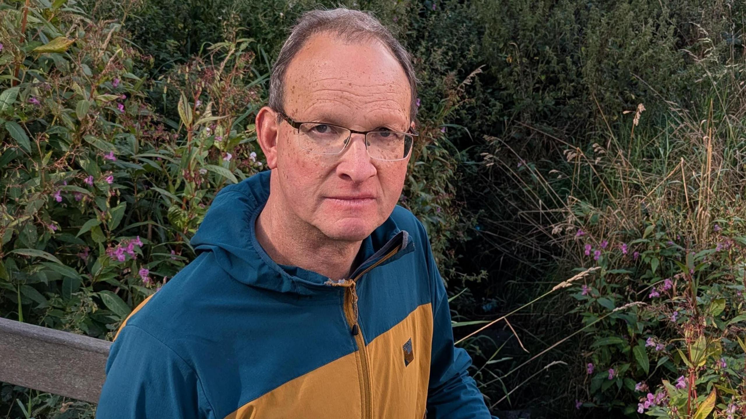 Councillor Peter Walton standing in front of flowers and bushes. He has short hair and is wearing glasses and a blue and brown outdoors-style jacket.
