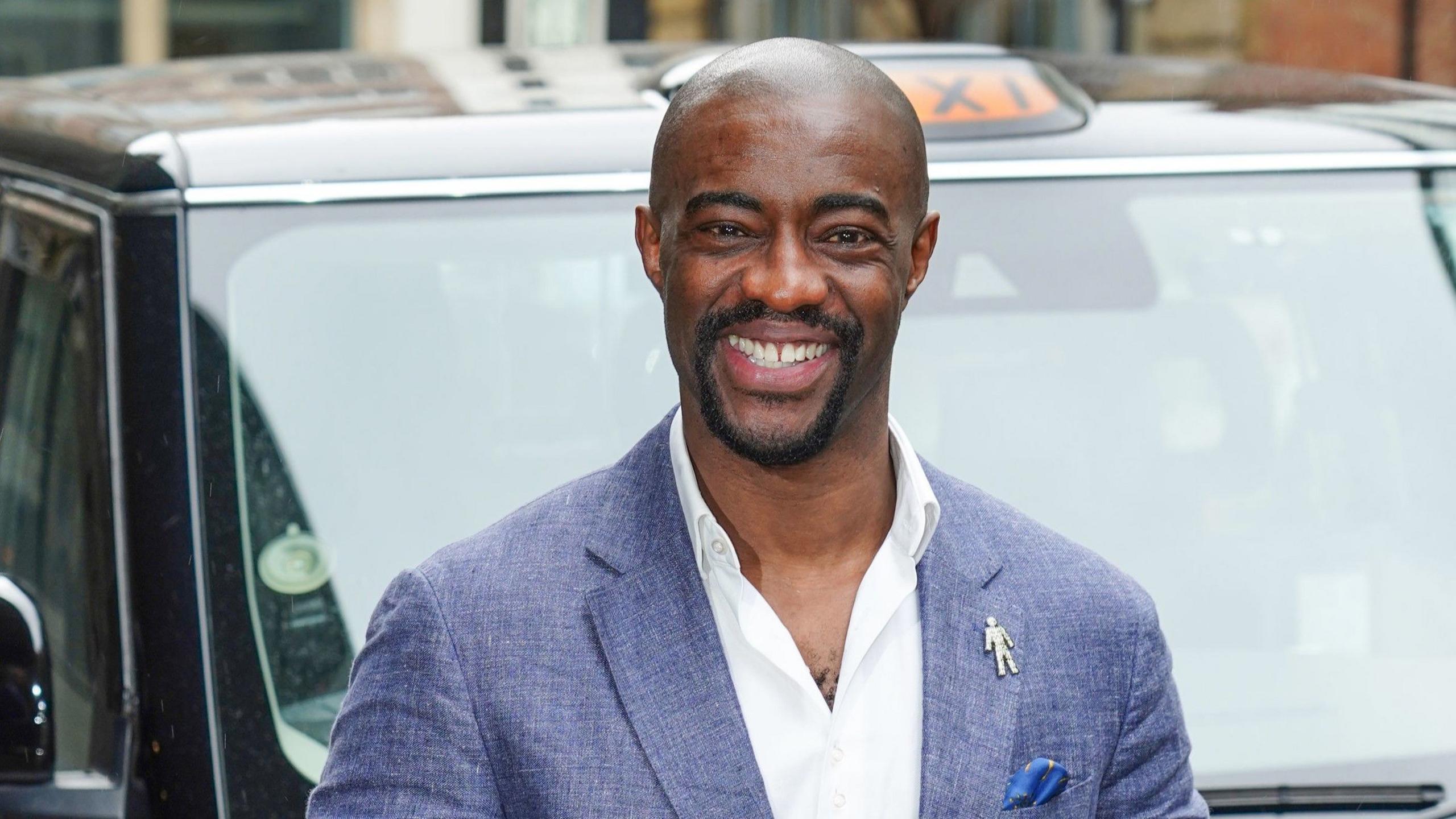 Tim Campbell stands in front of a black London taxi. He is wearing a mid-blue jacket and a white open-necked shirt. He has a bald head and a short black beard. He is smiling.