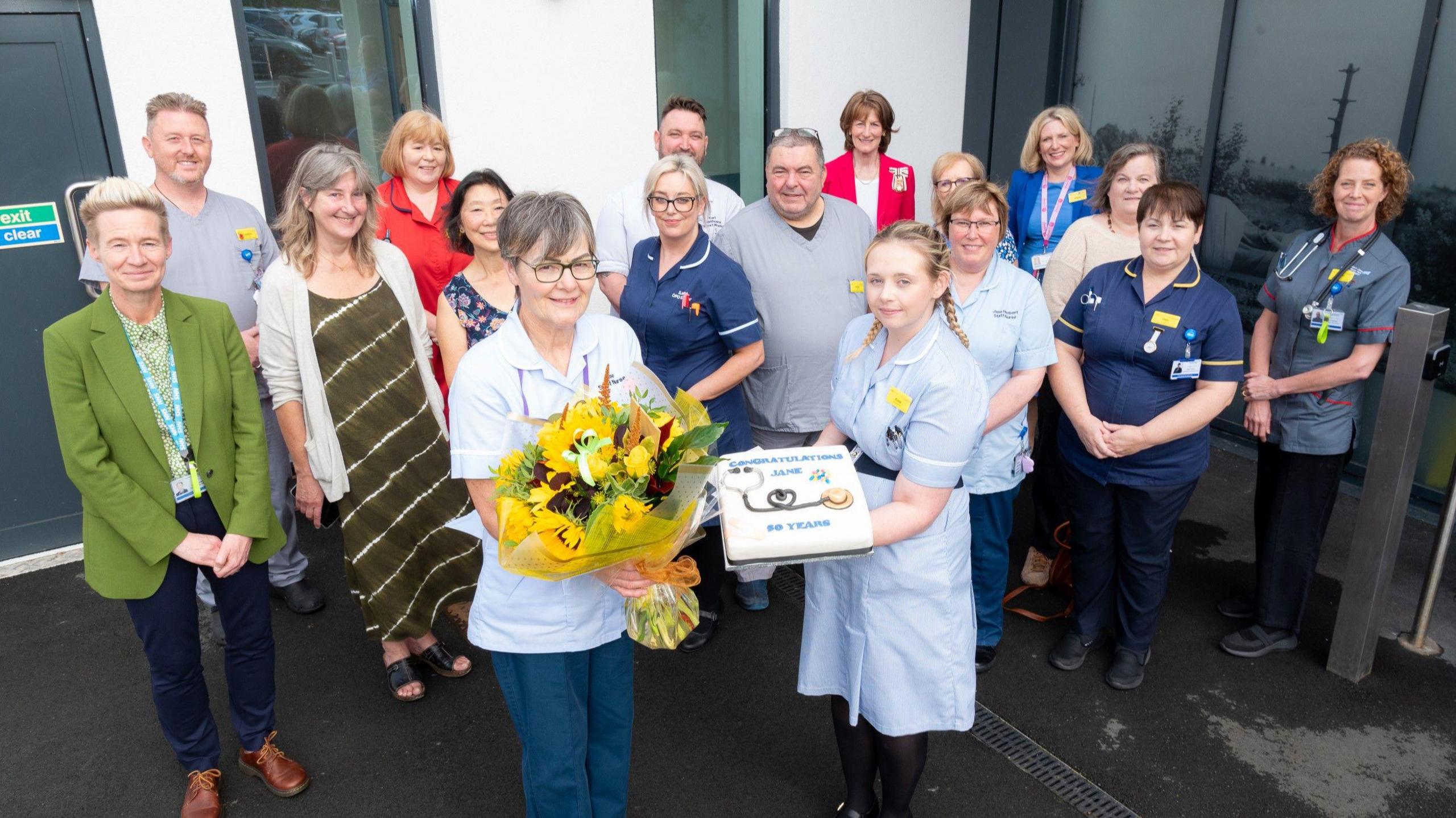 A group of 17 men and women stand facing towards the camera, some of them are wearing nurses uniforms. In the centre a woman holds a bouquet of yellow flowers whilst a woman next to her holds a cake