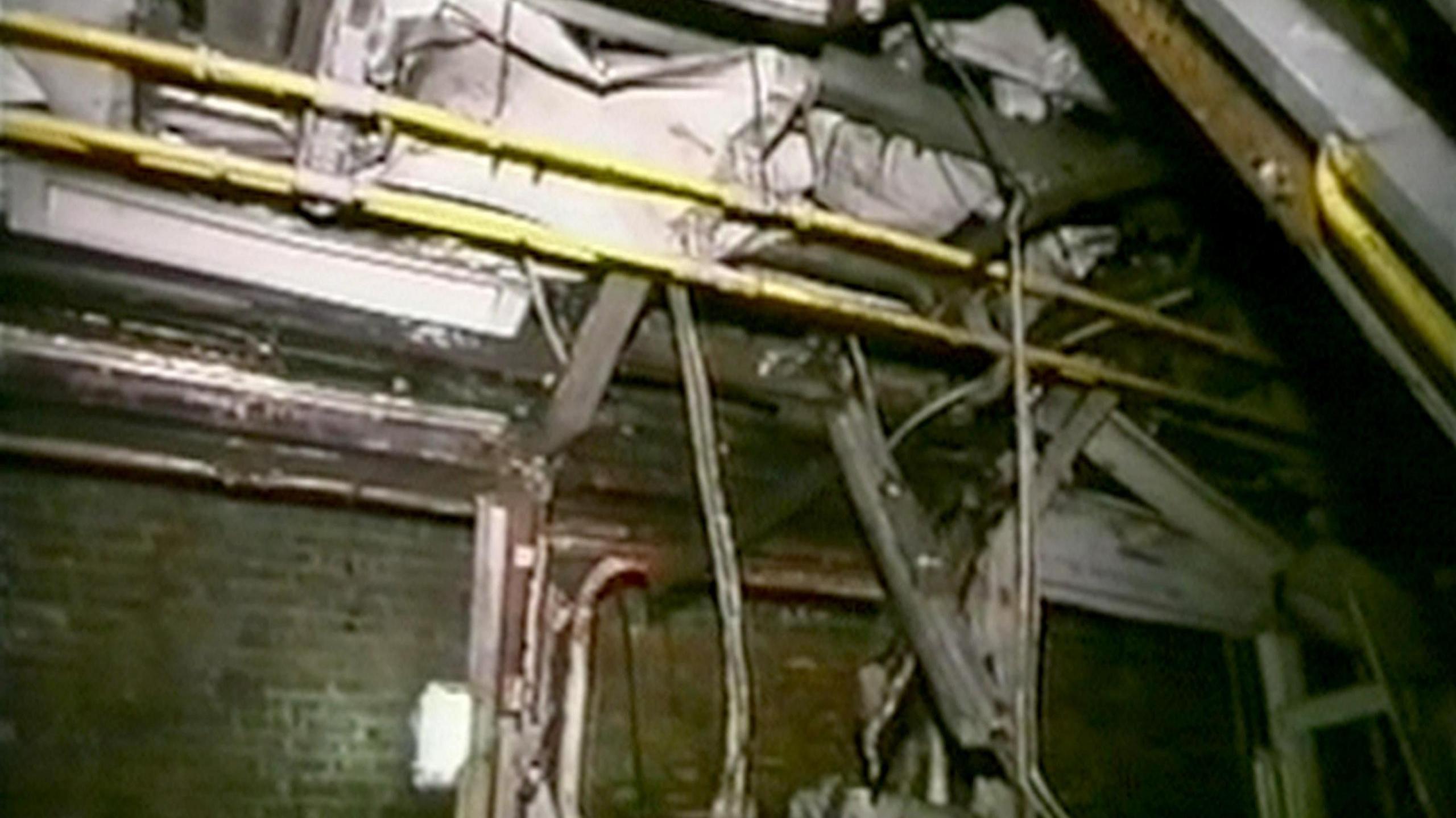 Wreckage hangs from the ceiling of a rube carriage. The yellow handrails are in tact across the ceiling, but debris is falling around it. In the background are the brick walls of the London Underground.