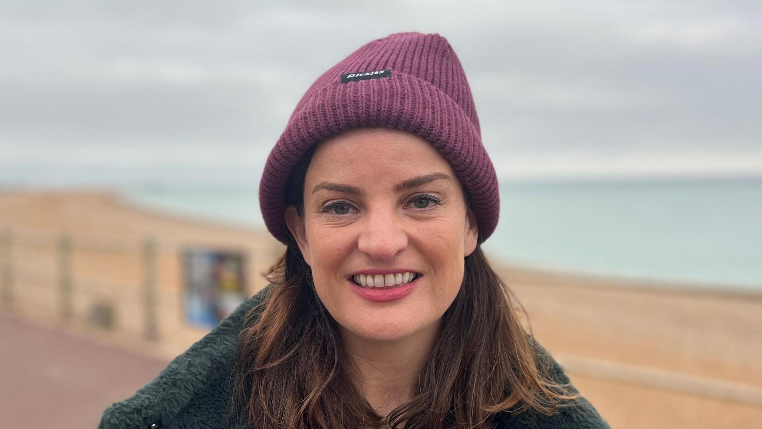 A woman with long brown hair looking and smiling at the camera. She is wearing a fark fluffy coat and a purple beanie. The beach and sea is behind her.