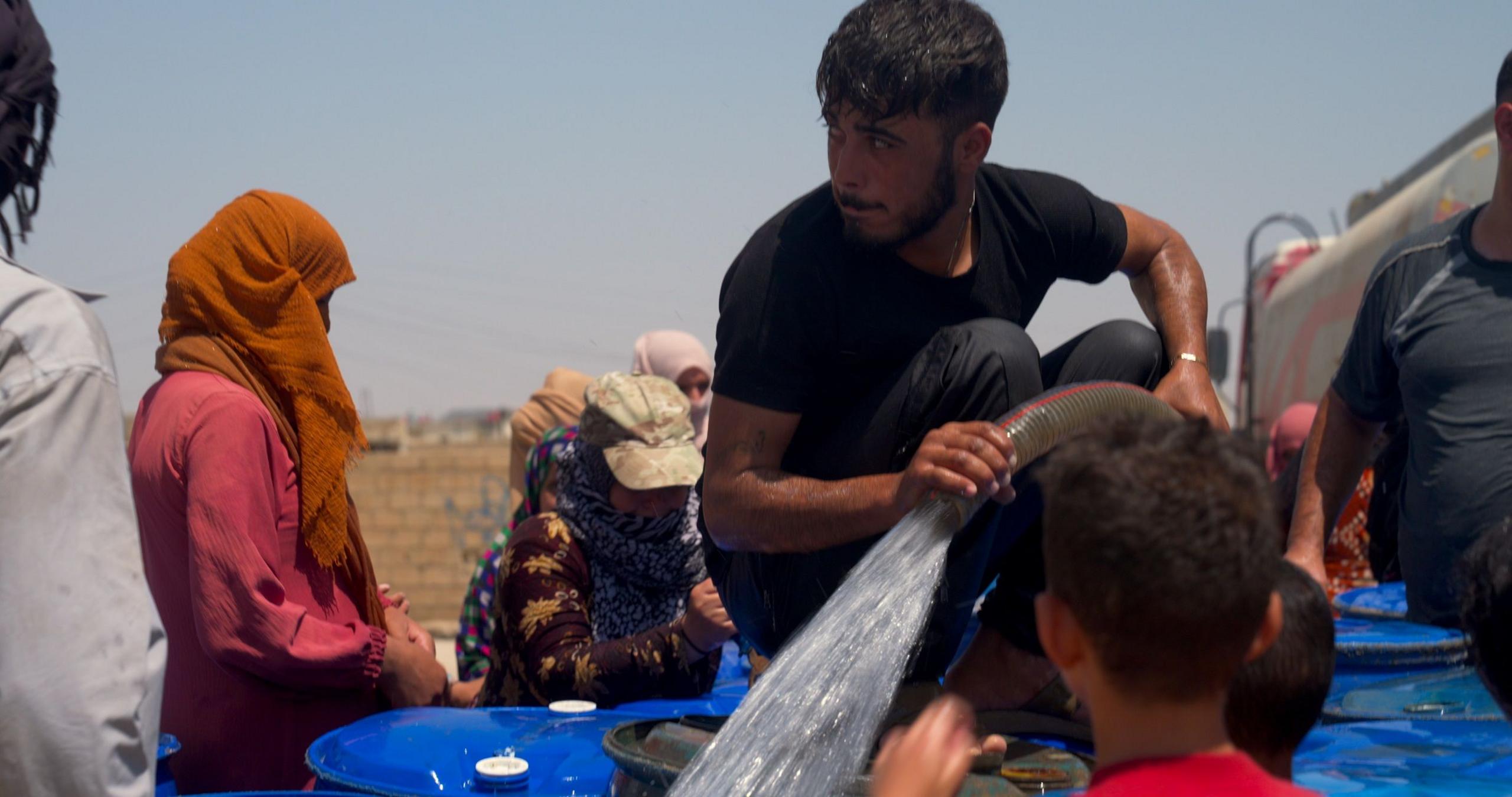 A man pumping out water into drums and barrels, with people waiting for water in the background