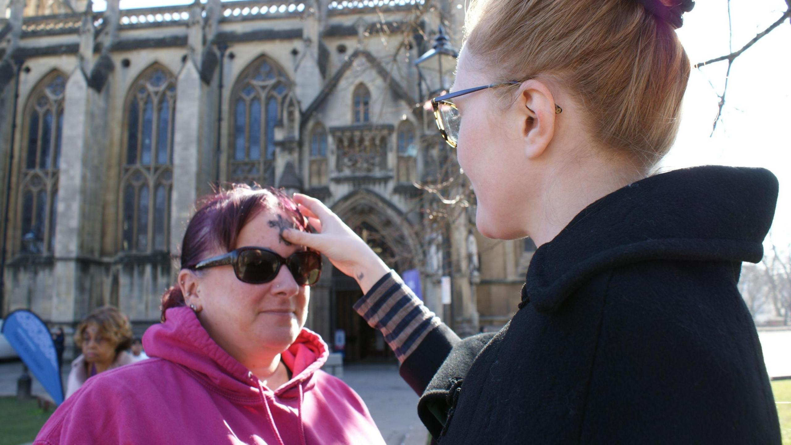 A young woman with tied-up hair applies ash to the forehead of another woman who is wearing a purple hoodie and dark glasses outside Bristol Cathedral on Ash Wednesday.