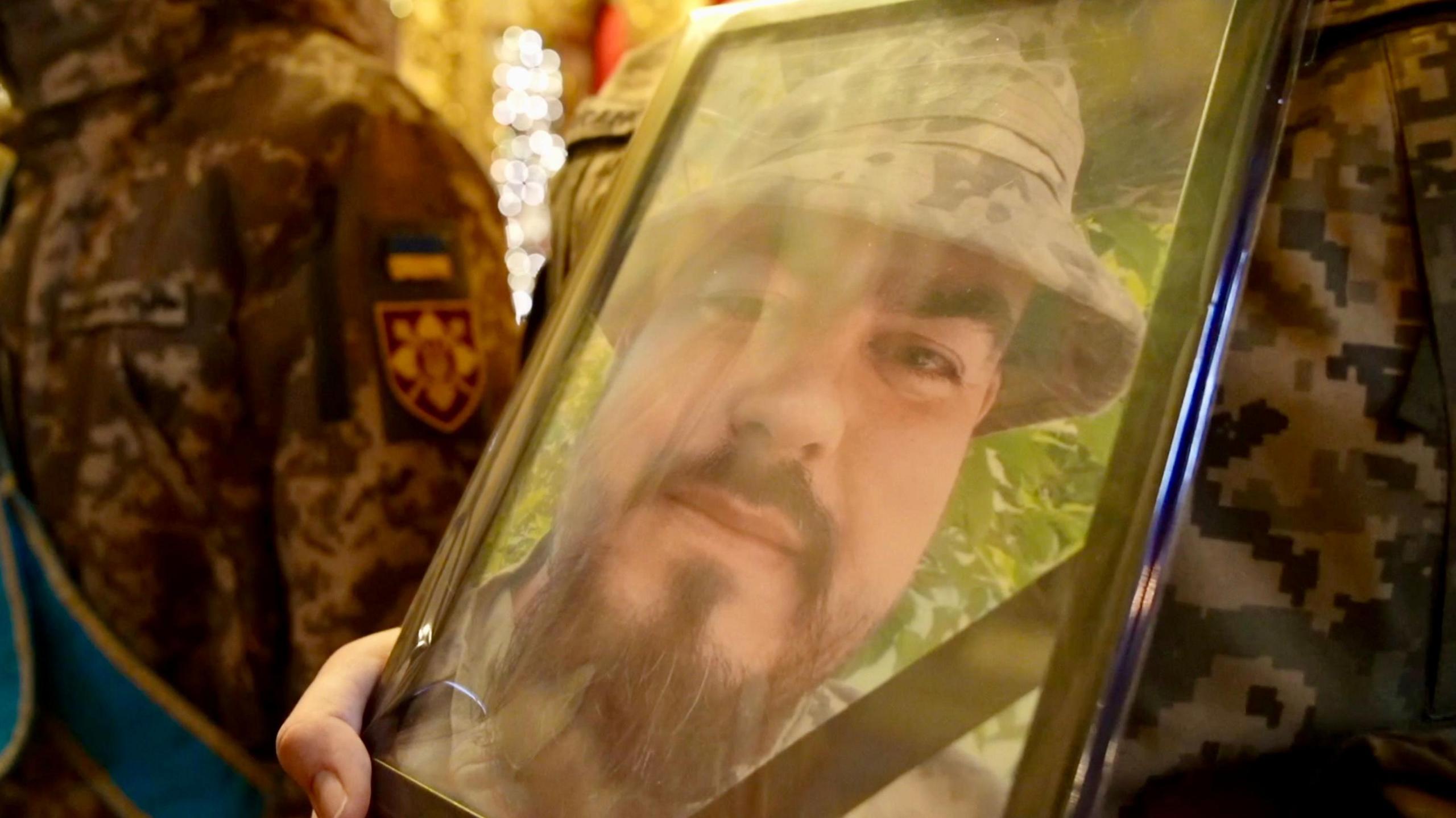 A soldier holds a framed photo showing Andriy Kusmenko smiling at the camera wearing a camouflage hat