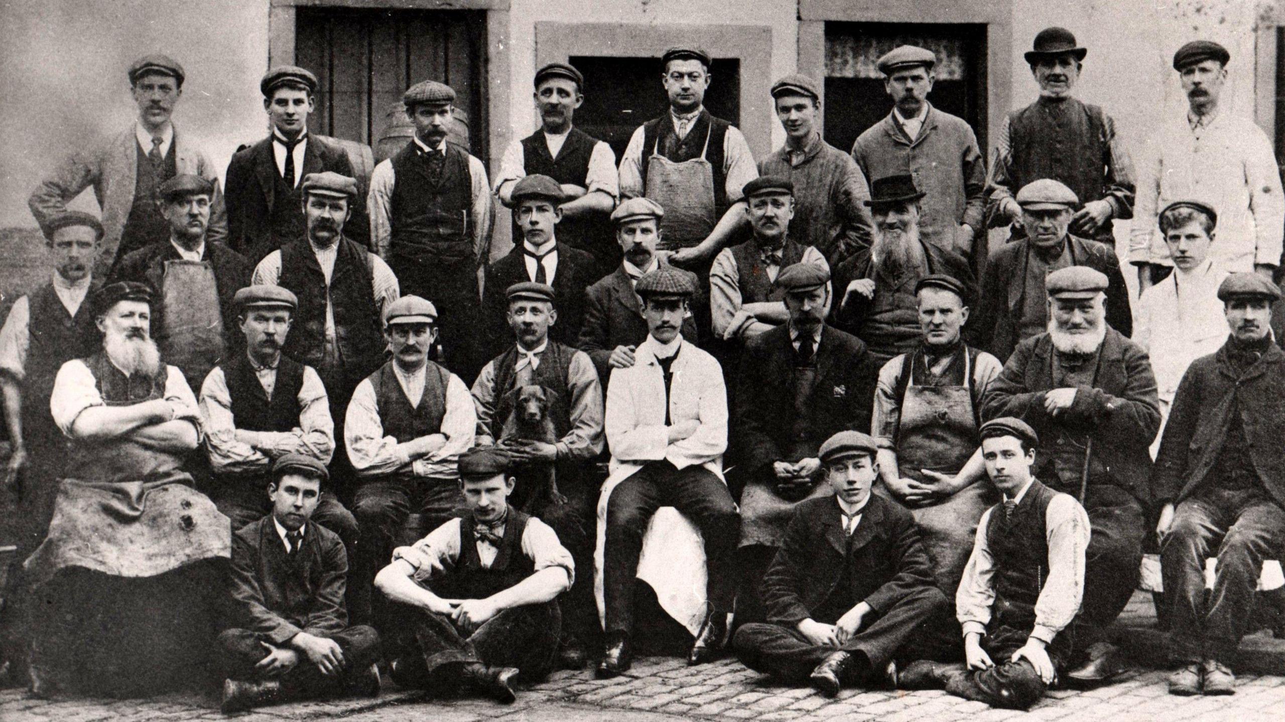 A black and white image of brewery workers from 1892. The men, of all age ranges, are in rows, some standing at the back and others sitting on the cobbles. They are all wearing caps bowler hats. 