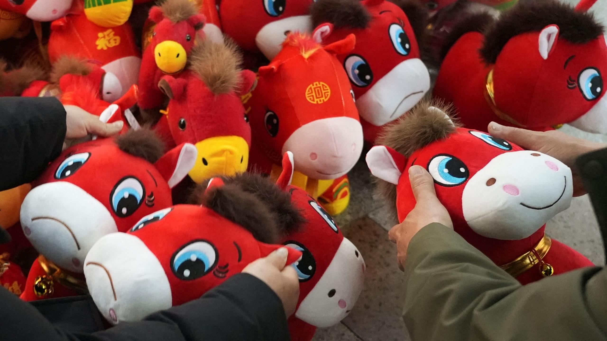 Customers look at crying horse and smiling horse plush toys ahead of the Chinese Lunar New Year. There are lots of different red plush horses. Some are smiling and some are sad. People are holding up different ones
