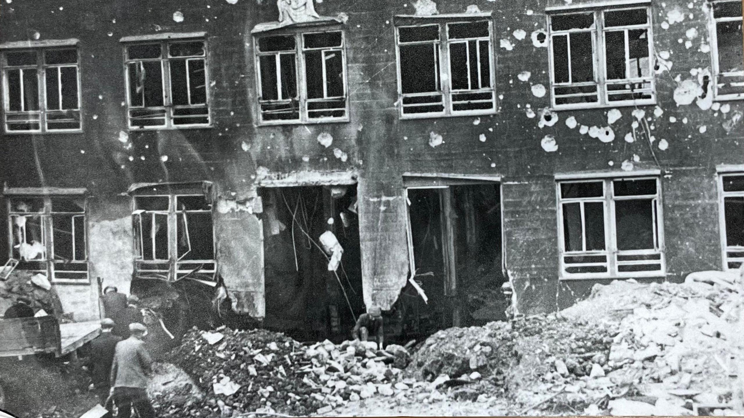 A black and white photograph of a building damaged by bombing. There is debris on the ground and holes in the walls.