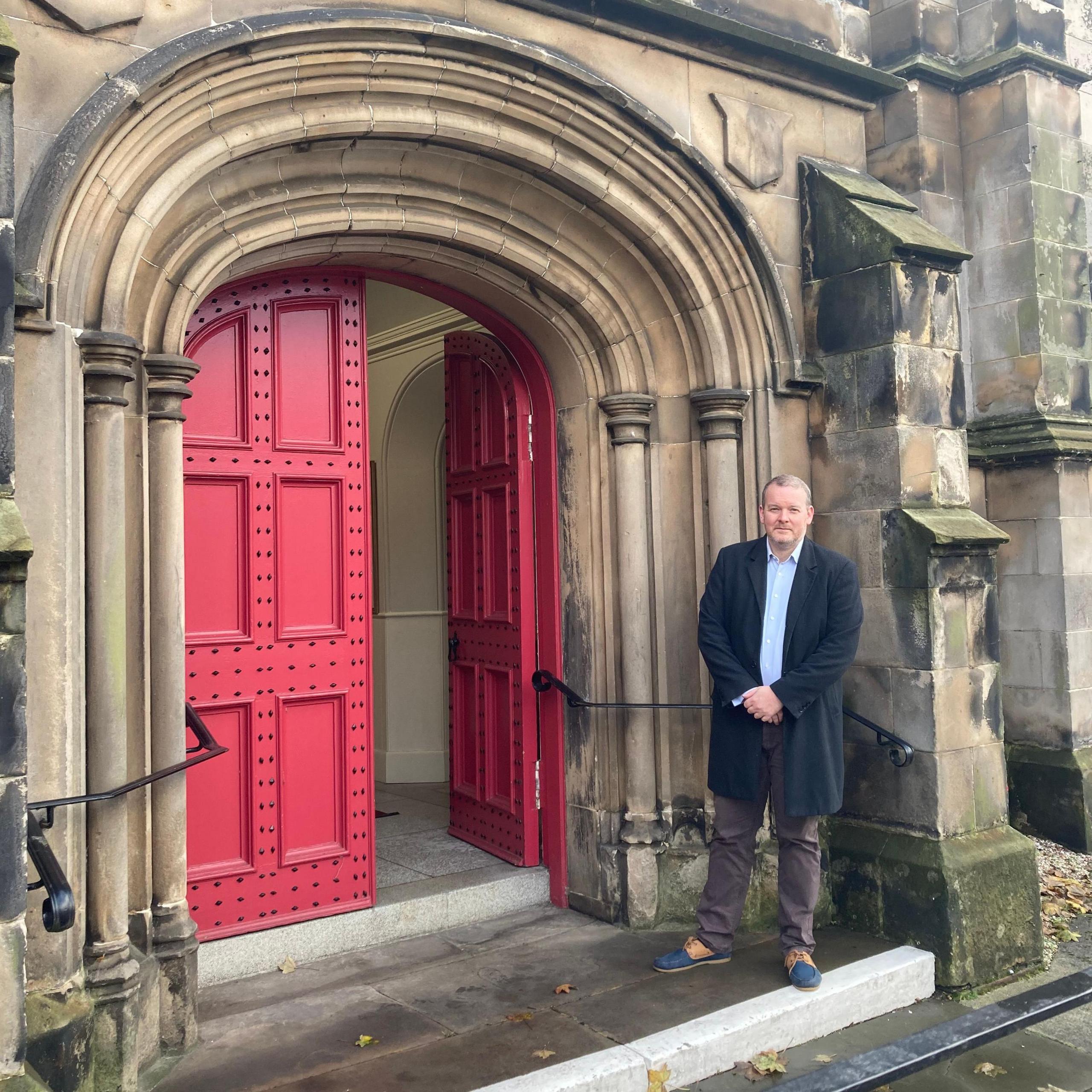 Brunton Theatre Trust chairman Michael Stitt standing in front of the red doors of the North Esk Church. He is wearing a light blue shirt under a dark blue overcoat, dark trousers and blue shoes and is standing on a white step outside the church. Red doors are in a rounded alcove with a black handrail on the side and light stonework.