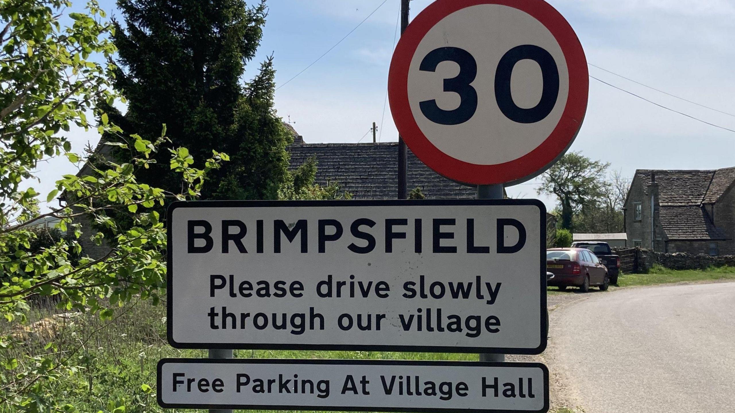A photo showing a black and white welcome to Brimpsfield sign, with a 30 miles per hour sign above it. In the background is a road, car and a house