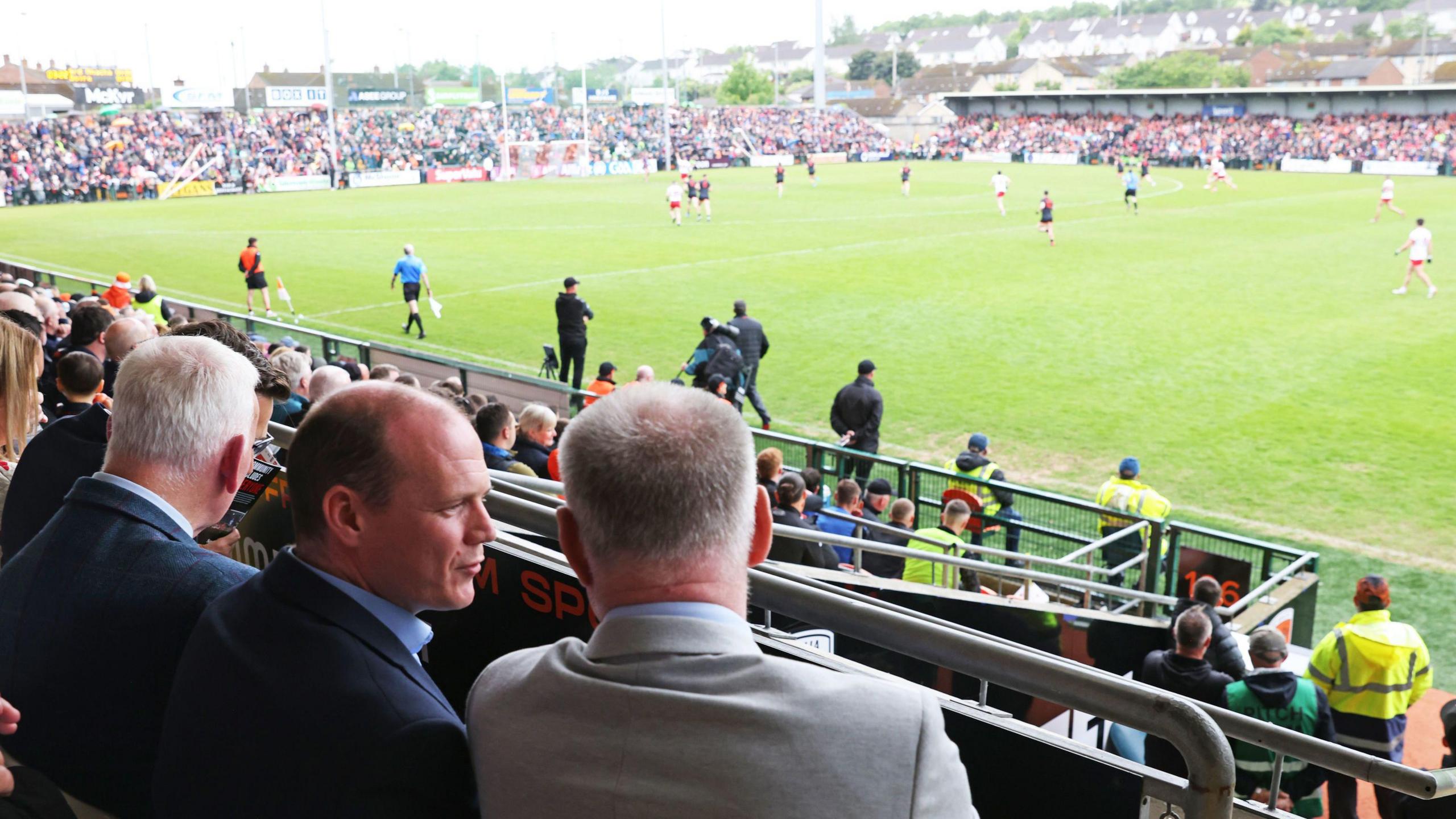 Gordon Lyons is seen from the back in the stands at the stadium. The players are on the field in front of him. He has his head turned in conversation with a man, who we know is Ulster GAA secretary Brian McAvoy.