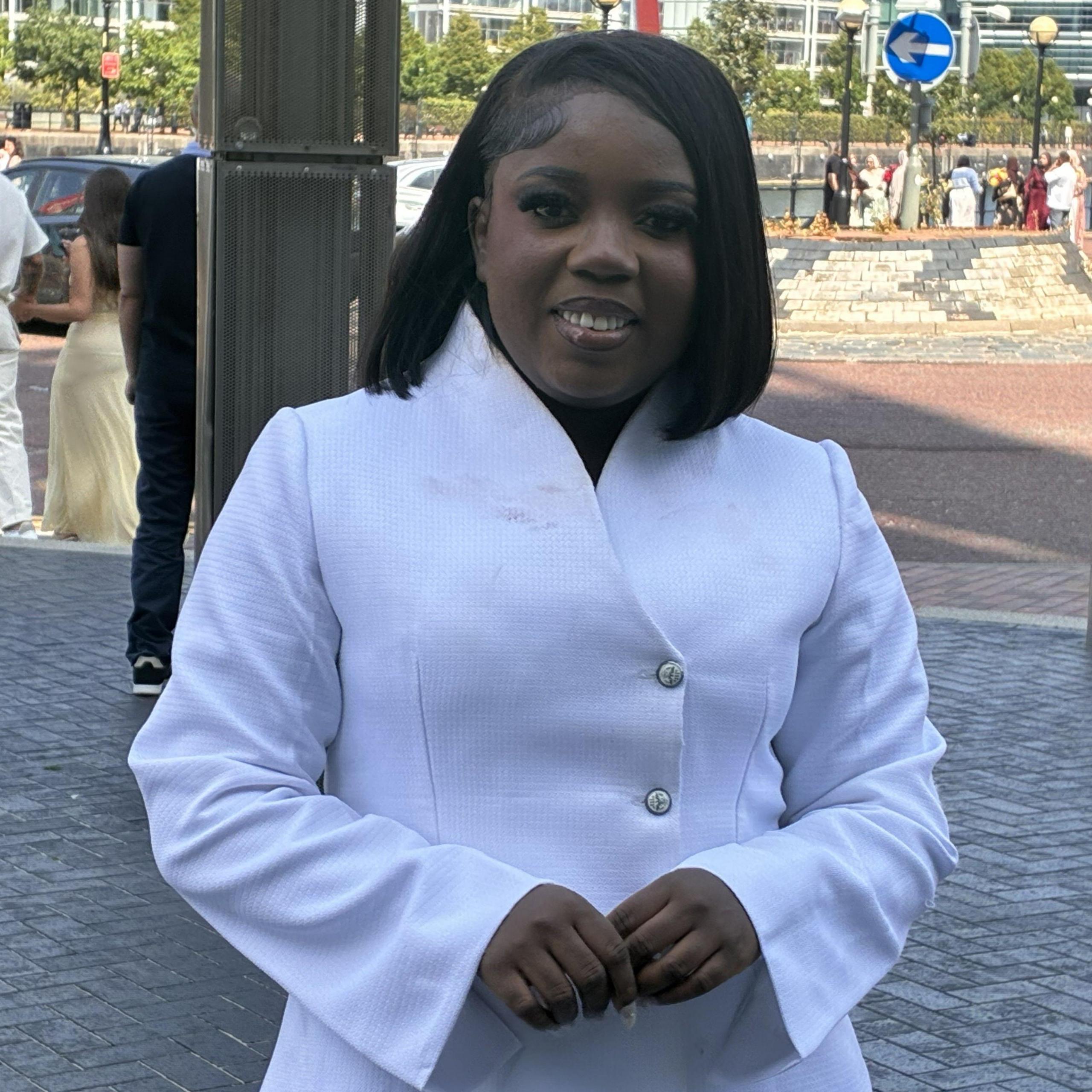 A woman stands in a white suit jacket with a raised collar and flared sleeves. Buttons run down the front slightly off centre. Behind her are several tall apartment buildings.