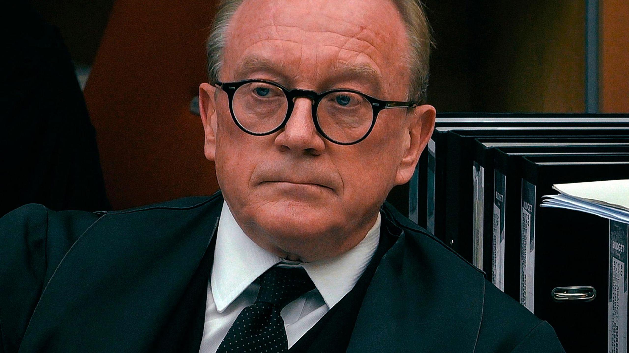 A photo of prosecution lawyer Alex Prentice in his office. He wears a black suit jacket with a very formal white shirt and black tie. He has short hair and glasses with round rims. Behind me are black folders of court documents. 