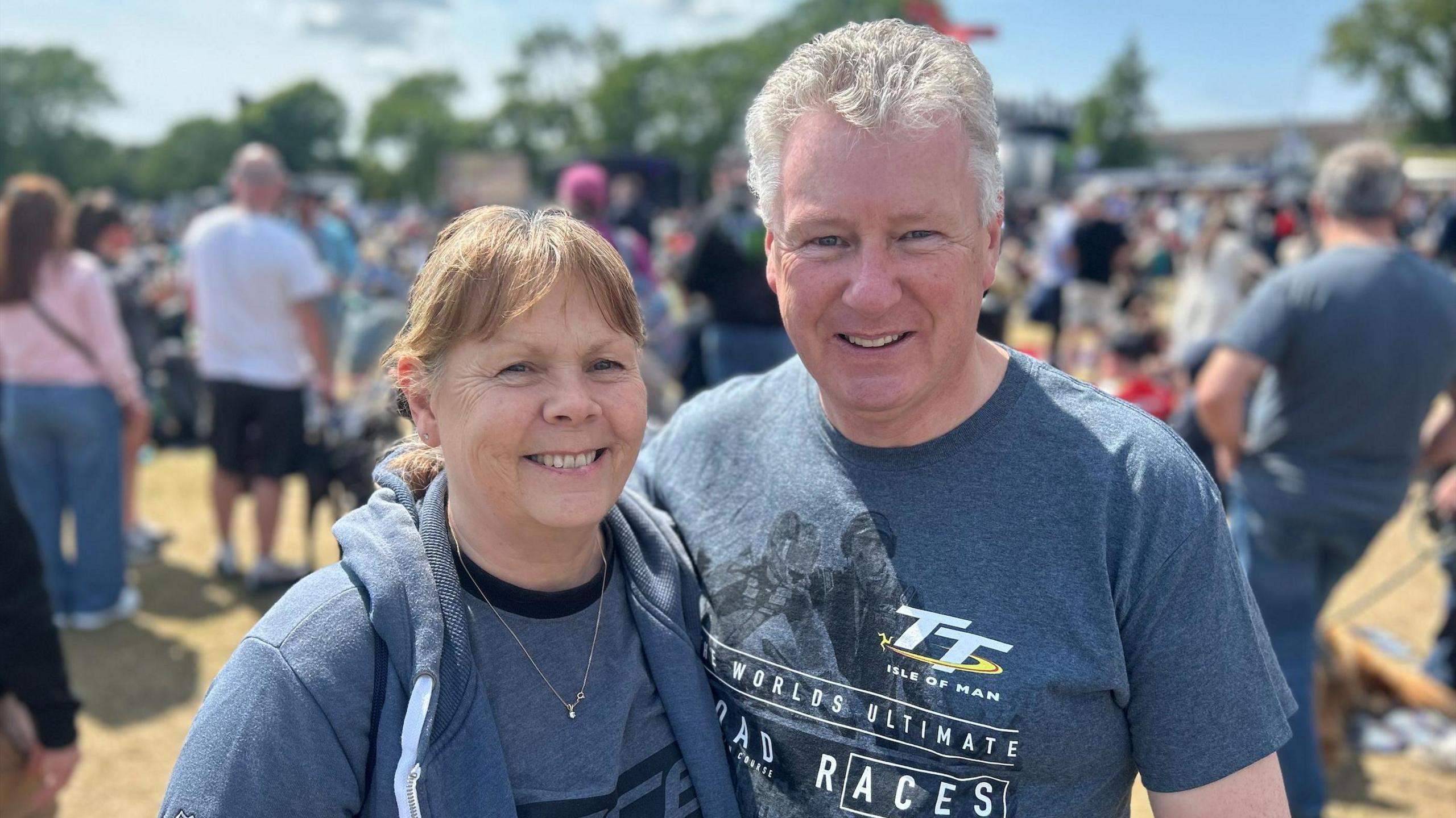 Ali and Rob Graham smile, they wear grey TT t-shirts and stand in front of a crowd. Ali has brown hair in a pony tail and Rob has short grey hair.