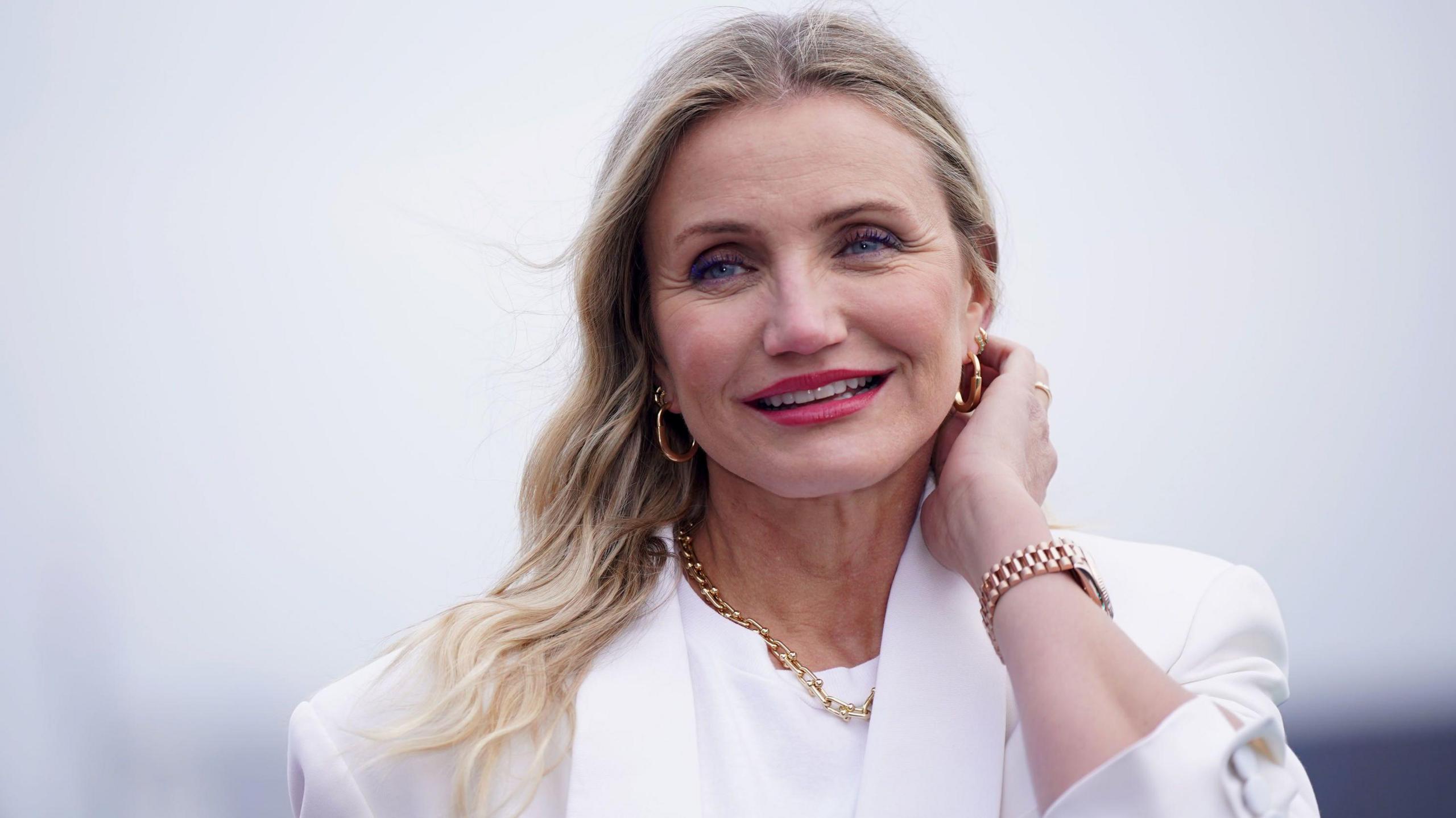 Cameron Diaz wearing a white blazer and white blouse. Her hair is open and over her right shoulder, and she wears gold hoop earrings and a gold chain necklace.  With her left hand, she is pushing hair behind her ear.