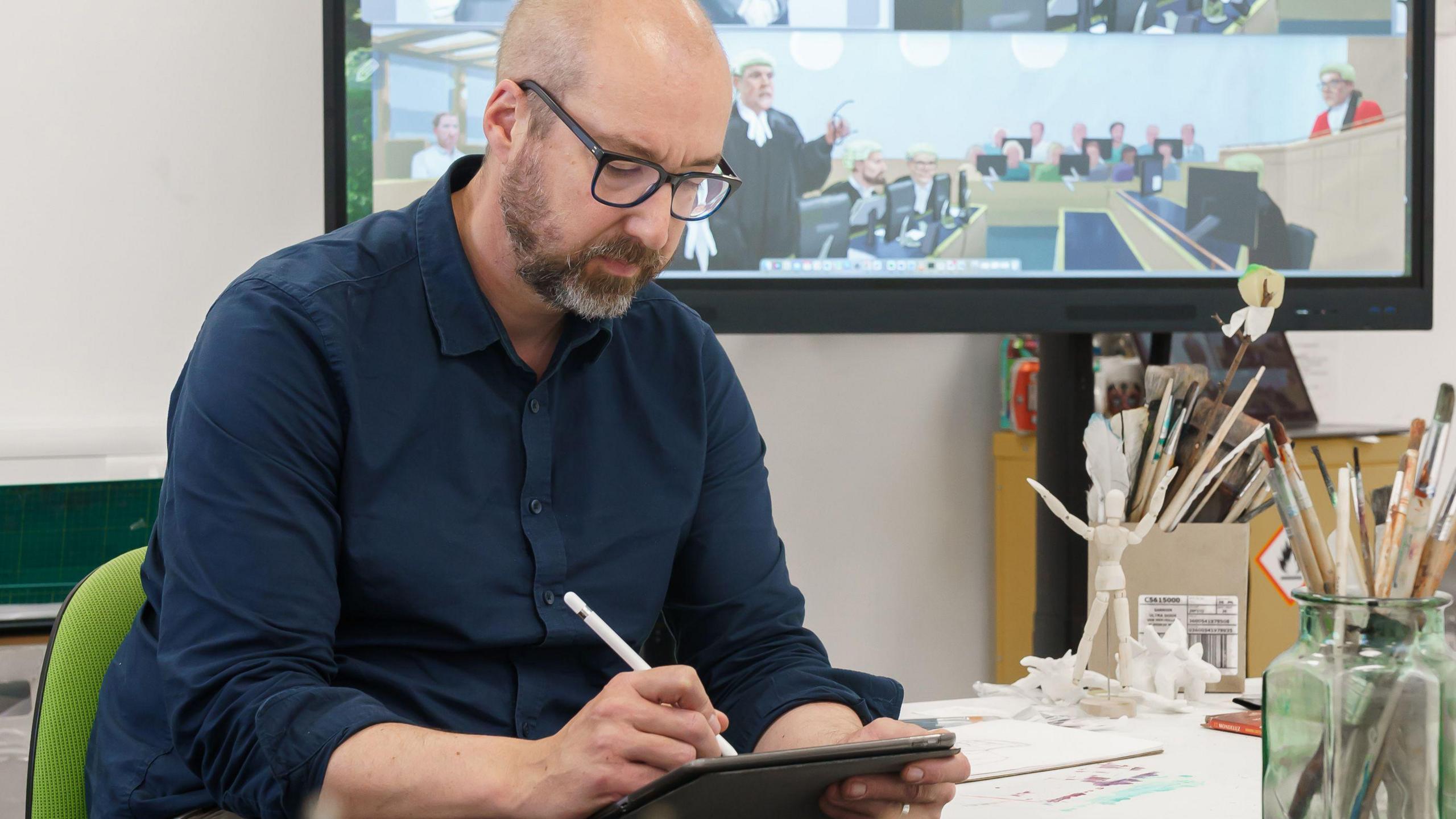 Nick Lewis sketches on his iPad using a white stylus pencil. He is wearing a navy shirt and glasses and is bald with a dark and grey beard. Behind him is a TV screen showing one of his sketches form the trial, which depicts a barrister standing and speaking to a judge.