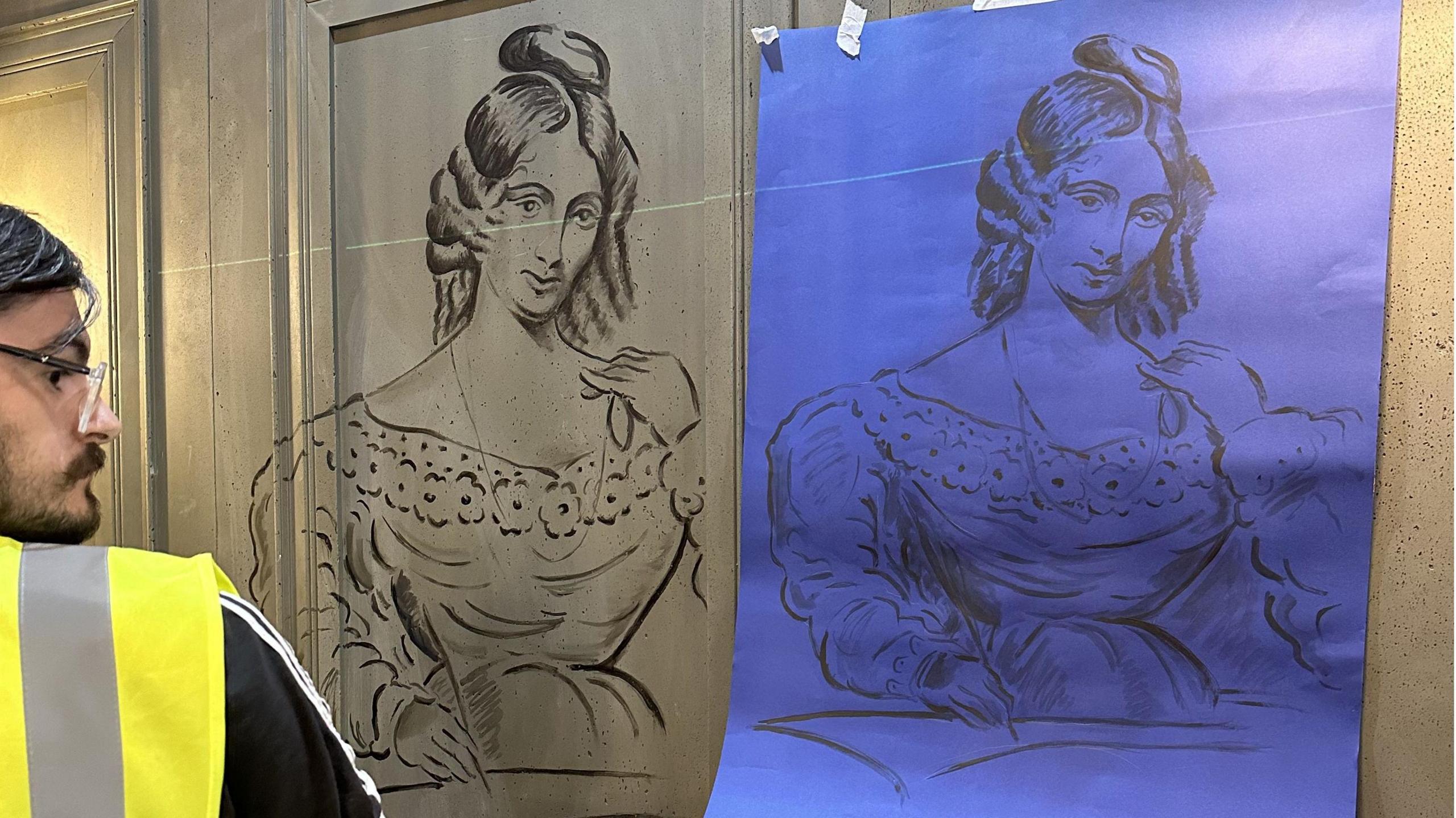 A man in a high-vis vest is using a line drawing reference image of Mary Shelley to paint her portrait onto a wall