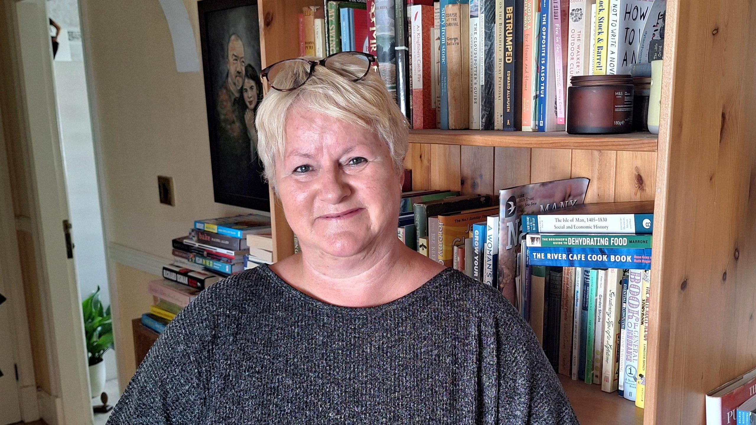 A close up of Sue King. She has short blonde hair and has a pair of dark-rimmed glasses resting on the top of her head. Wearing a dark grey top, she is smileing while standing in front of a book case.