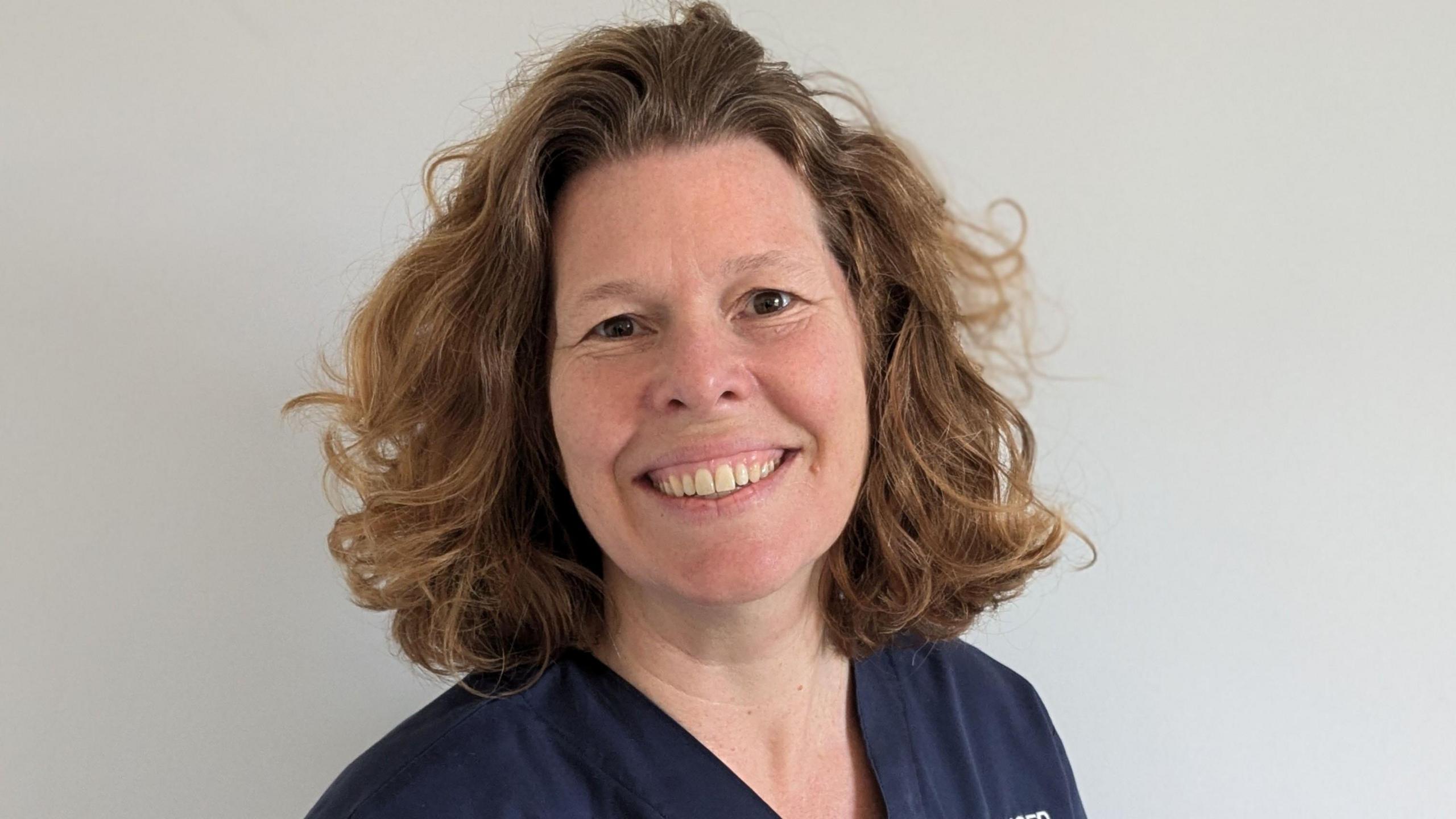 Hermione Bigwood smiles while wearing dark blue medical scrubs on. She has shoulder-length wavy hair which is light brown.