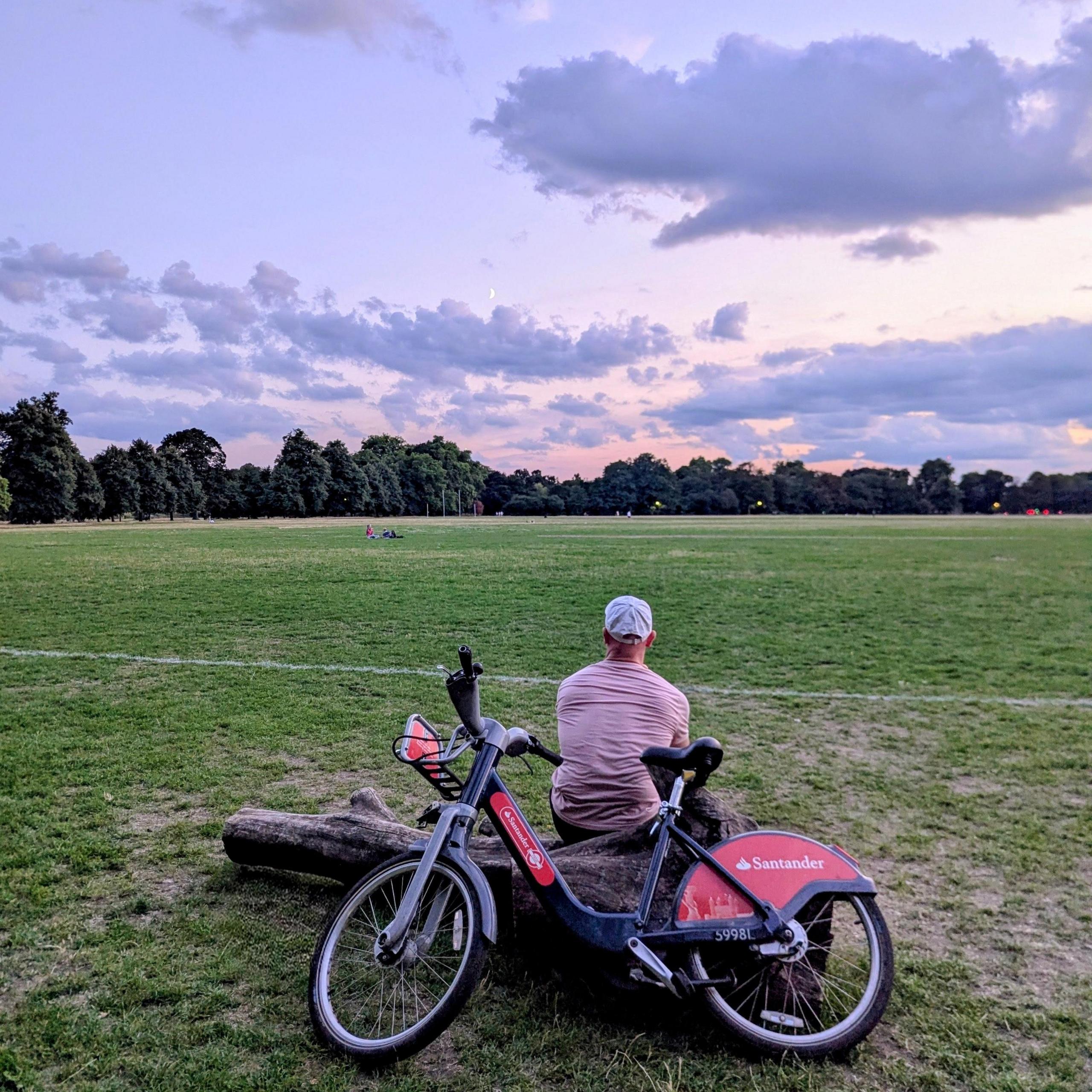 A man in a pale cap and pink T-shirt sits on a log in an open grassy park. A Santander Cycle is propped beside him. The sky above is filled with scattered clouds, and a line of trees is visible in the distance.