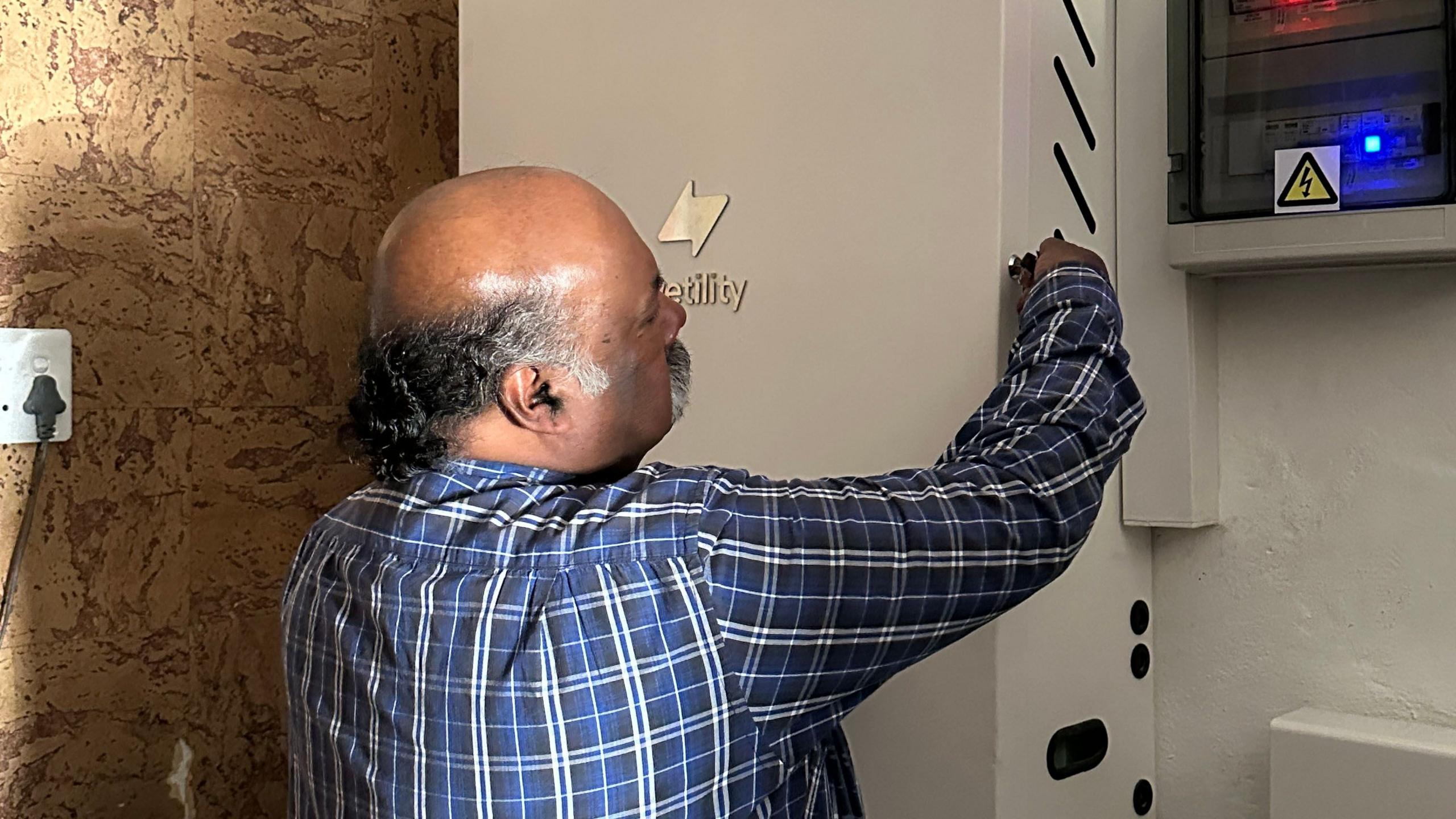 Mark Moodley, in a checked shirt, stands with his back to the camera fitting a key into a large meta box on the wall that contains the sola-power inverter and battery.