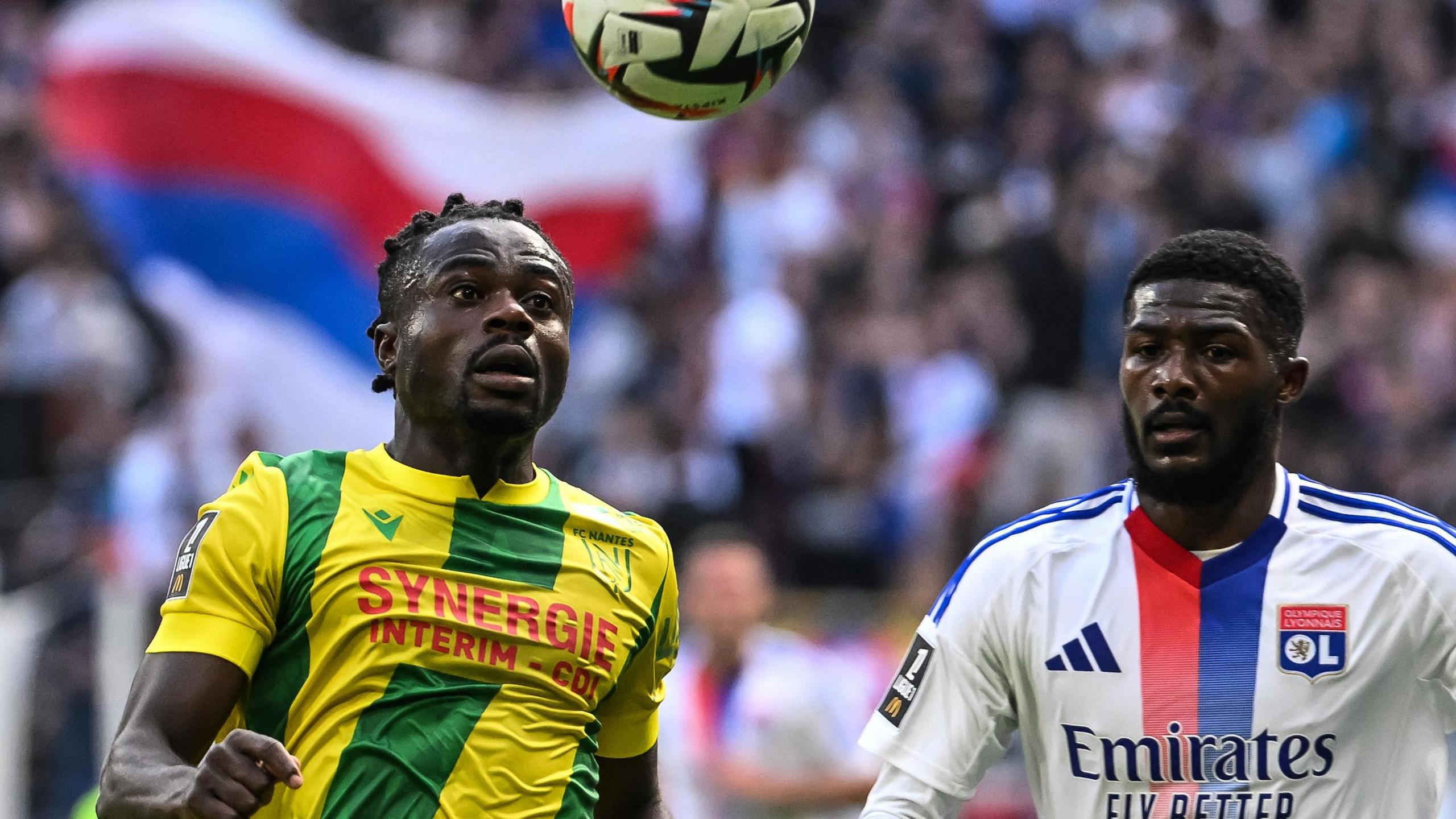 Moses Simon, wearing a yellow and green Nantes shirt, looks up towards a bouncing ball while Lyon player Ainsley Maitland-Niles is alongside him