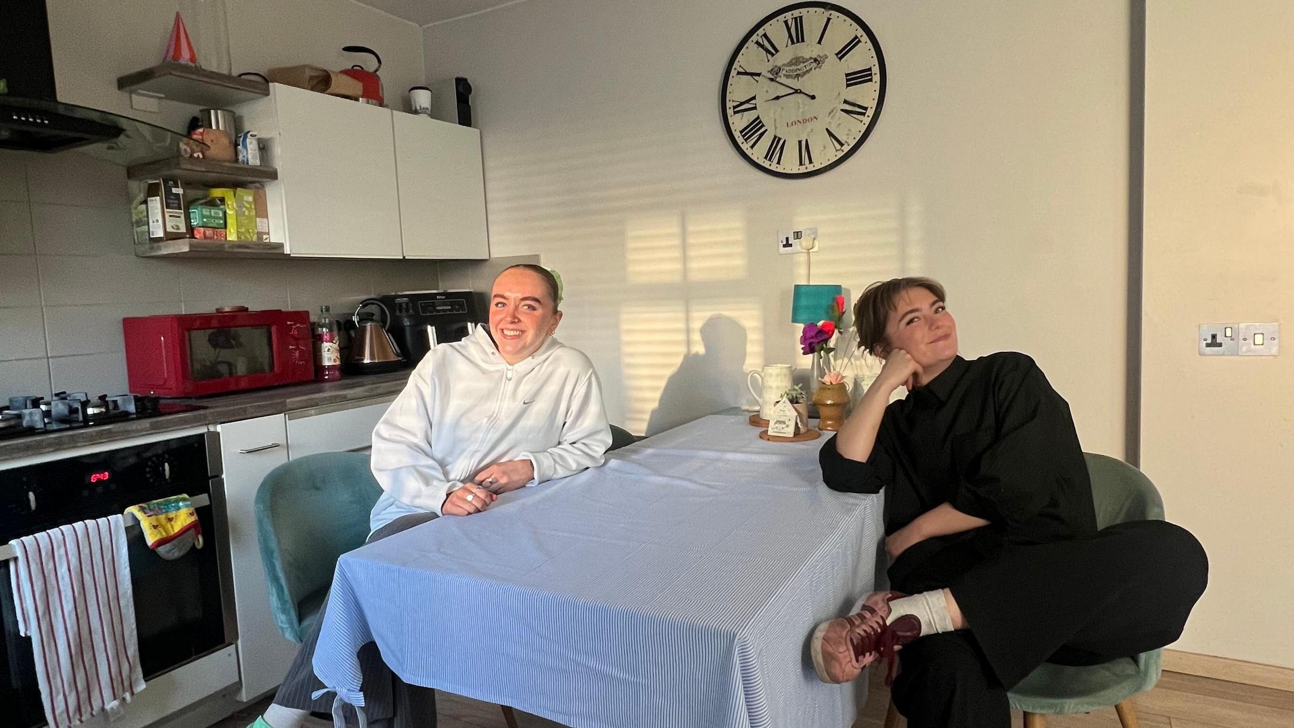 Ella Murray and her flatmate Maisy sit at a kitchen table which has a blue tablecloth. Both are smiling. The table is close to a set of kitchen cabinets and an over and hobs. There is a large, wall-mounted clock on one wall.