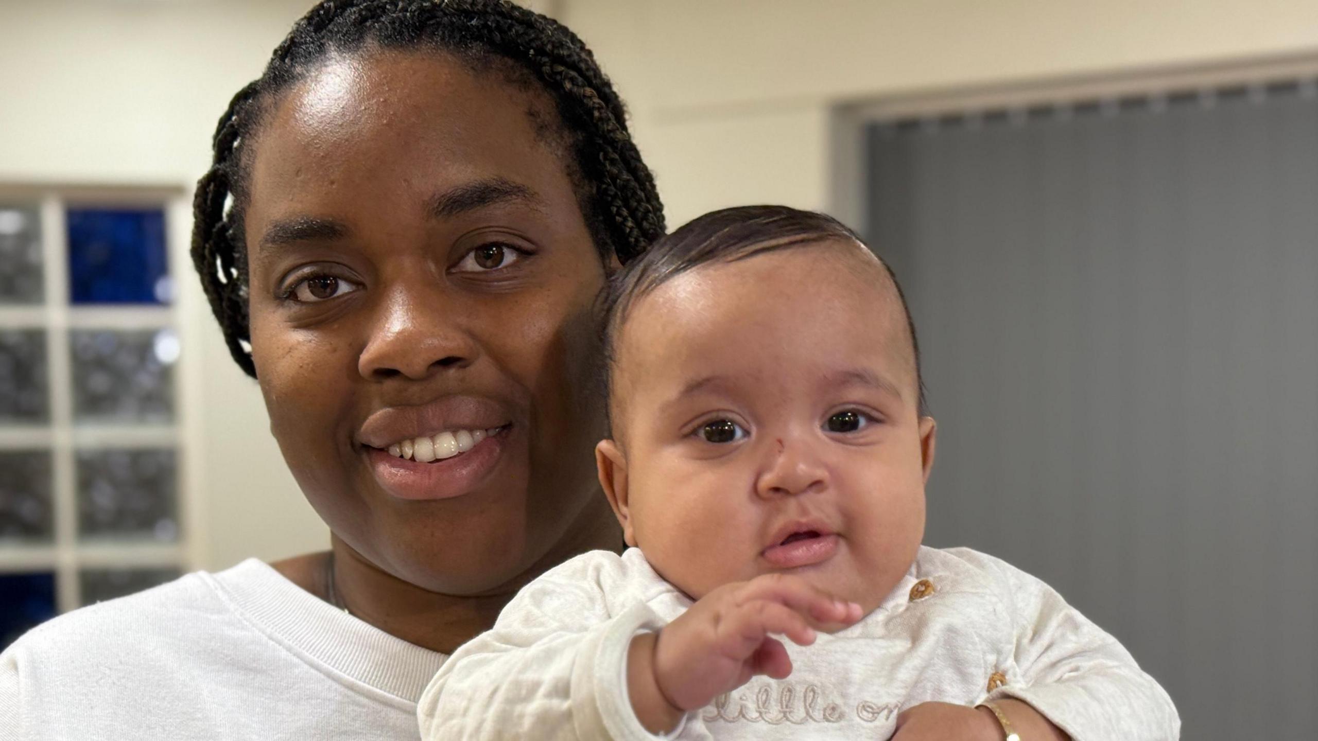 Sharna holds a child. She is in a white jumper and the child is in a cream-coloured baby suit with the words 'little one' embroidered onto it.