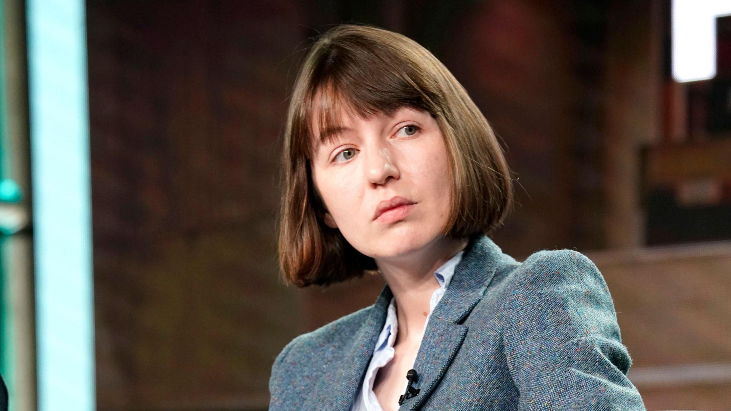 Sally Rooney, who has short brown hair and a fringe, and is wearing a woollen suit jacket and a shirt, listens to someone speak. 