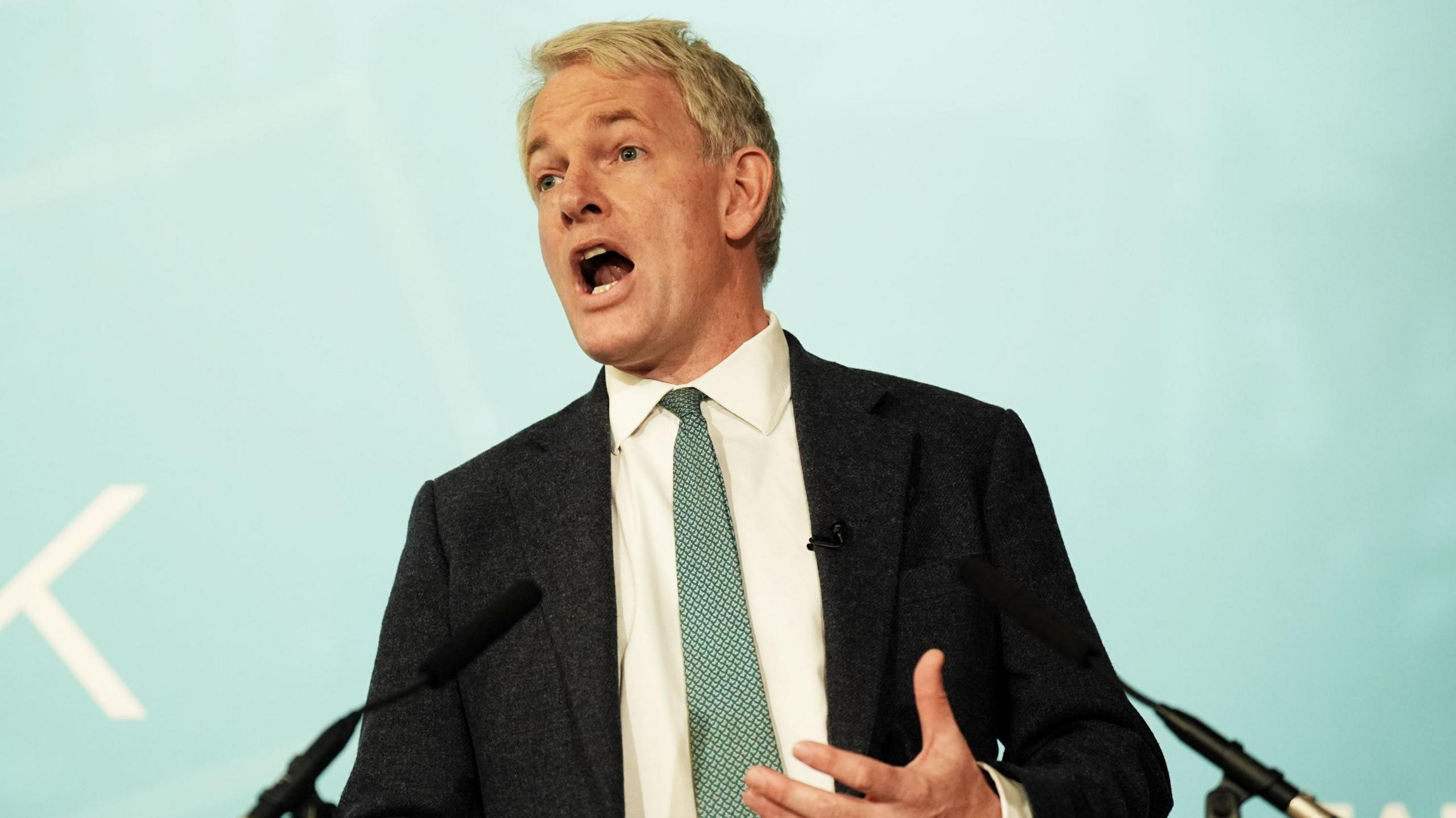 Danny Kruger talking at a lectern. He has a pale blue tie and a black jacket on, and is standing in front of a pale blue background.
