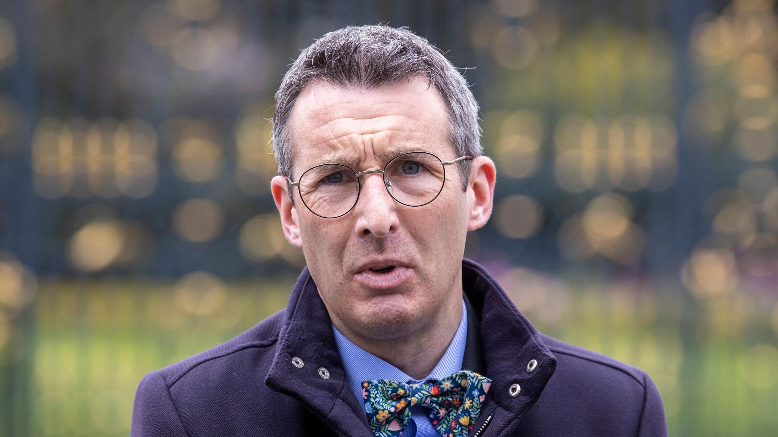 Andrew Muir has short grey hair and is wearing glasses. He is standing outside with a purple jacket and blue shirt on with a colourful patterned bow tie.