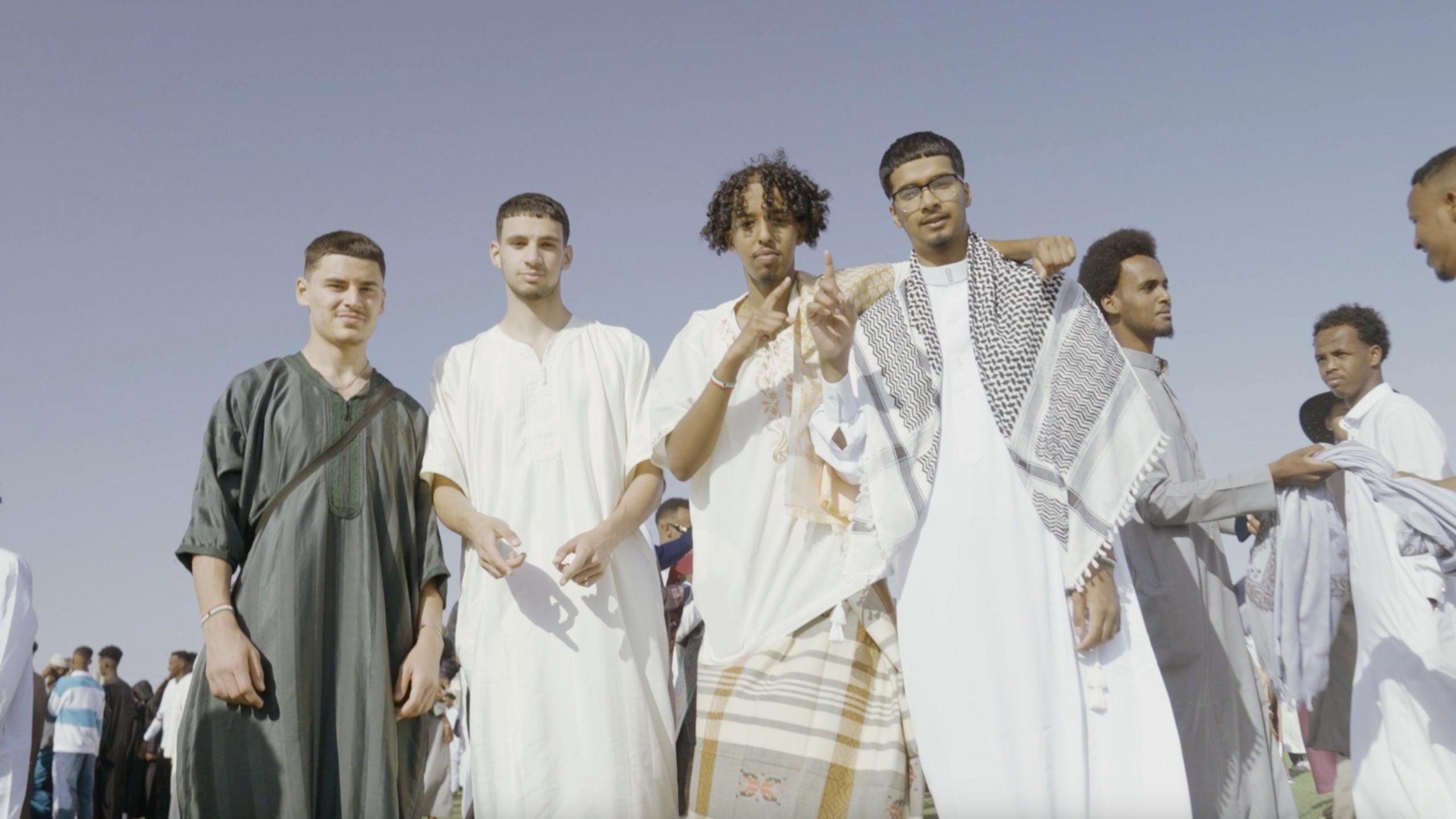 A picture of Zak Hajjaj, Anas Omar, Abu Finiin, and Kayum Miah standing in a crowd of people dressed in a mix of Muslim and Somali cultural attire.