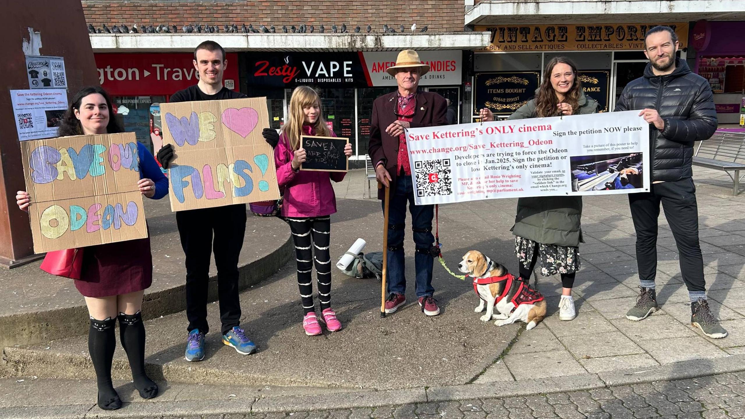 Six people and a dog standing in Kettering town centre holding placards. A woman is holding one which reads "Save our Odeon". Another says "We love films!" The dog is a beagle