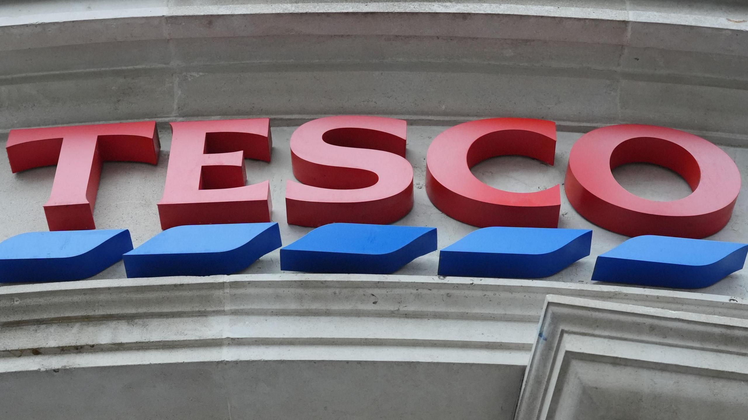 The logo of Tesco as seen on the front of a store. Large red font with blue dashes underlining it.